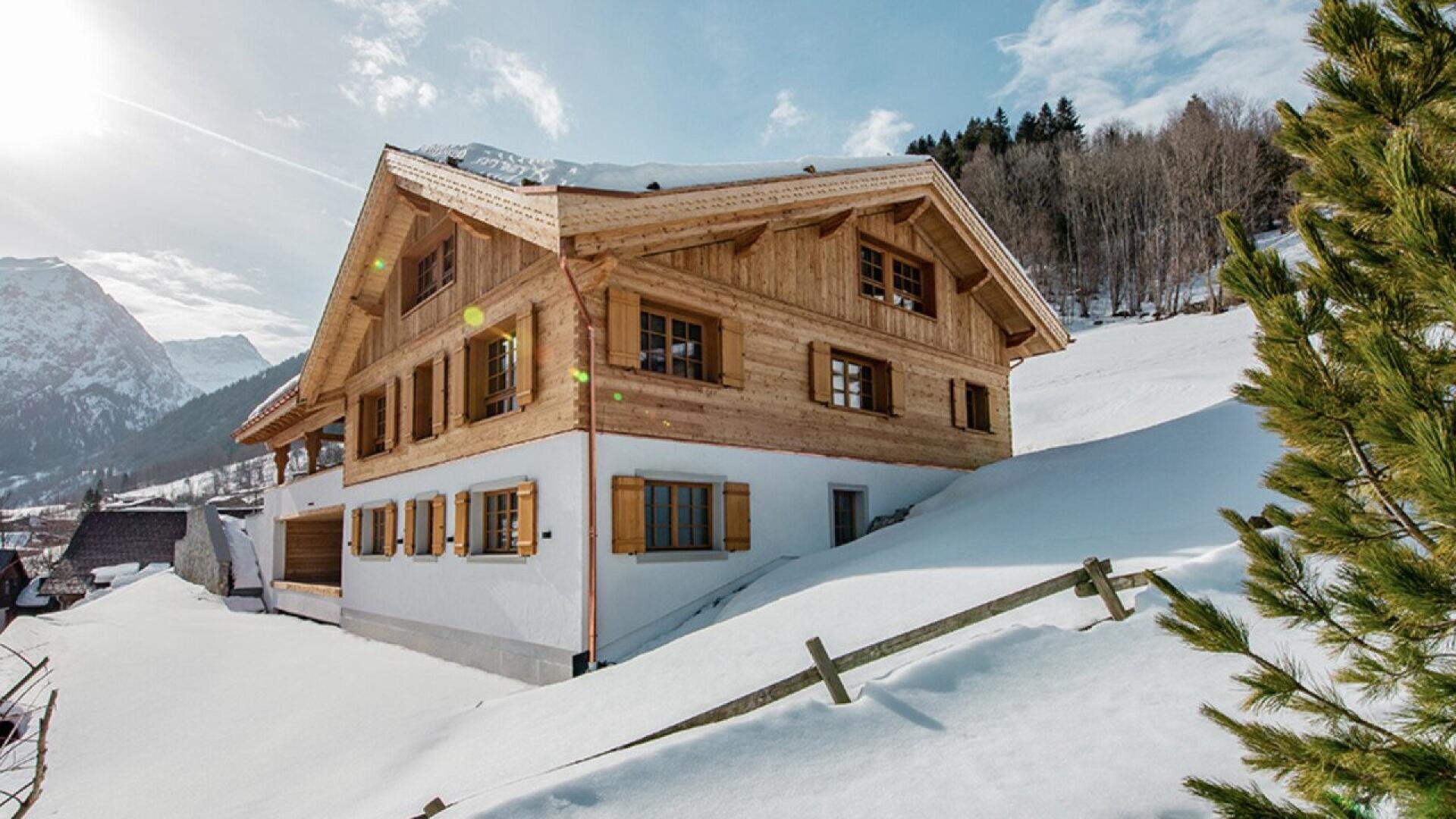 A wooden chalet-style house with snow-covered roof and ground, surrounded by mountains, pine trees, and a bright winter sky.