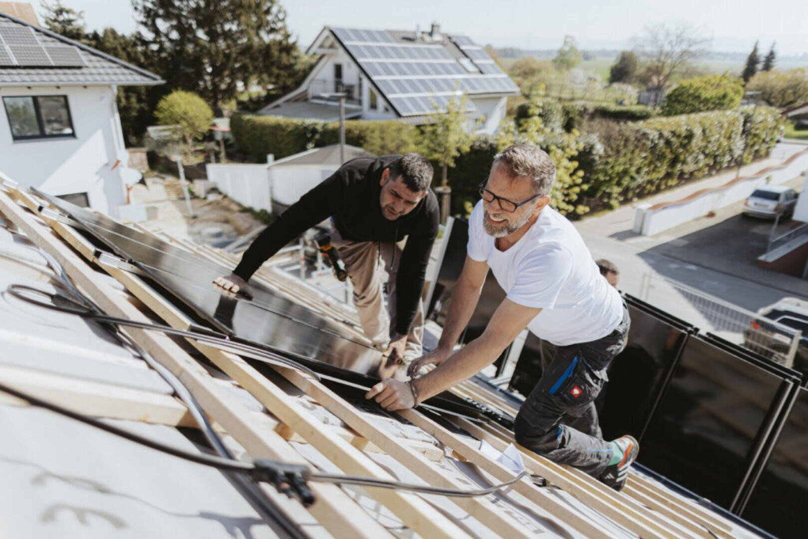 Two men are installing solar panels on a sloping rooftop in a residential neighborhood on a sunny day. They are working with tools and seem to be concentrating on their work, while solar panels can be seen on the roofs nearby.