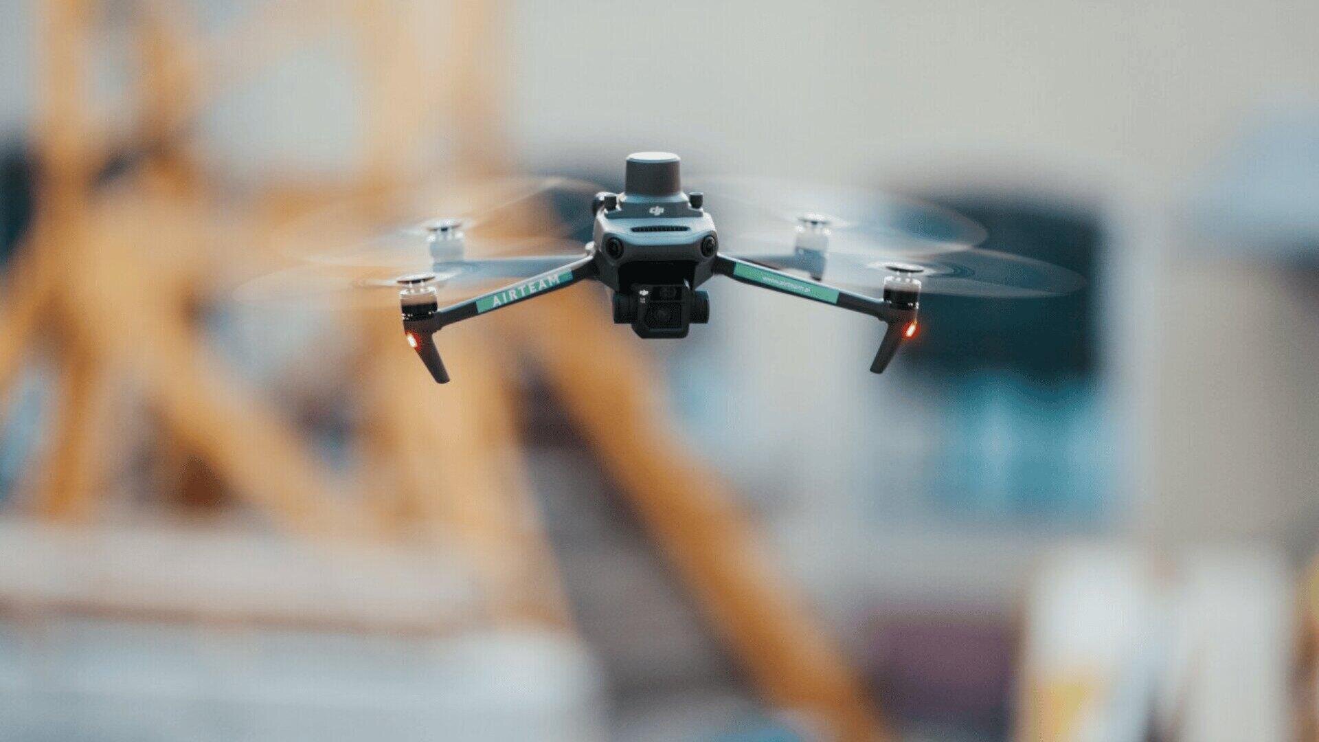 A black quadcopter drone with a camera hovers in midair, its propellers spinning and lights illuminated, with a blurred industrial background.