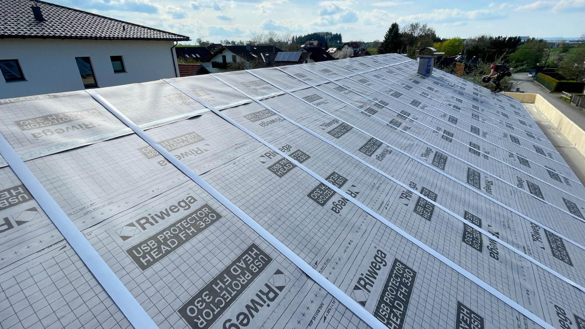 A large sloped roof is covered with Riwega waterproofing membrane sheets, with two workers installing them. Nearby houses and trees are visible in the background under a partly cloudy sky.