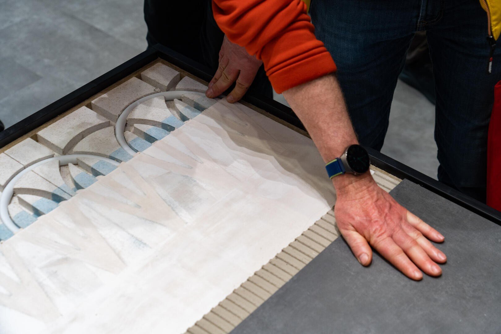 A person wearing a smartwatch and an orange case adjusts decorative tiles on a surface with both hands by arranging intricate patterns next to solid-colored tiles.