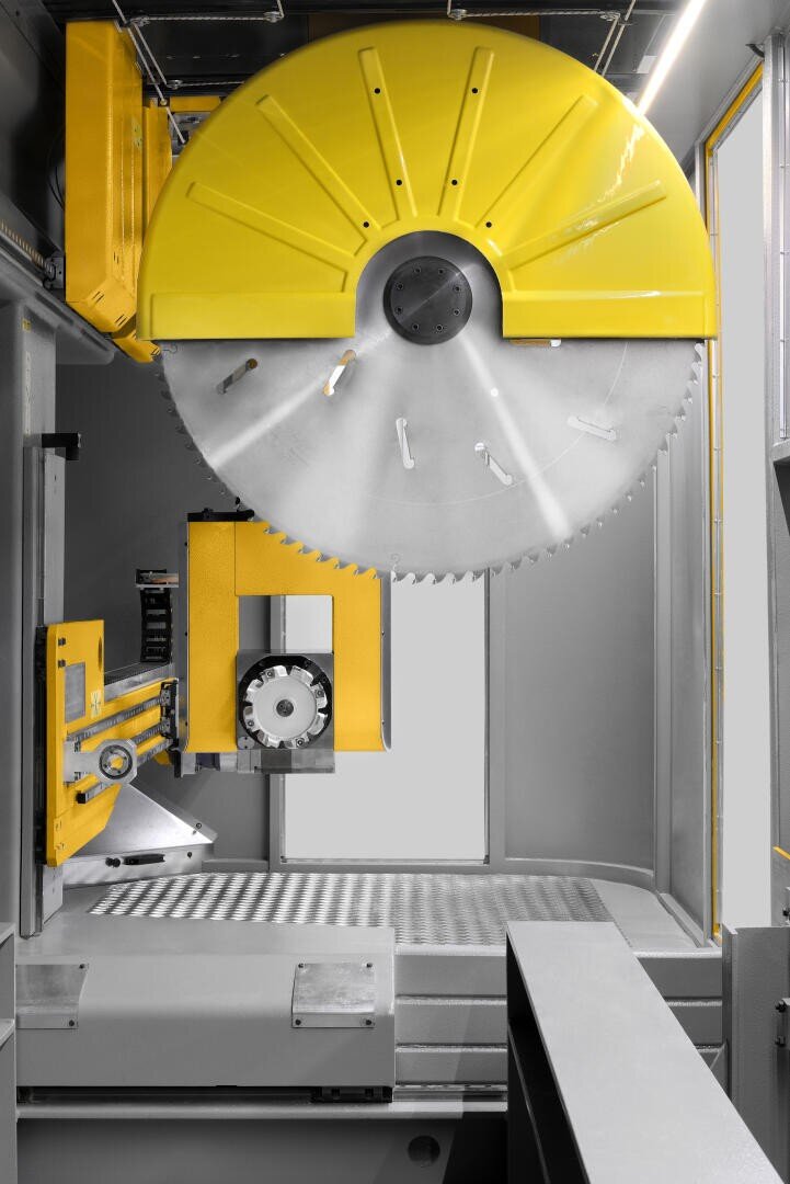 Eine große Industriesäge mit gelber Schutzhaube und silbernem Sägeblatt steht vertikal in einer Fabrikhalle. Gelbe Maschinenteile und Metalloberflächen dominieren die moderne Werkstattumgebung.