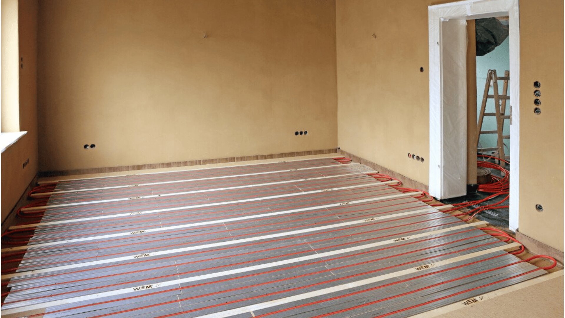 A room with bare walls and no furniture, featuring underfloor heating installation with visible red pipes and metal panels across the floor. A door opens to another room where a ladder is visible.