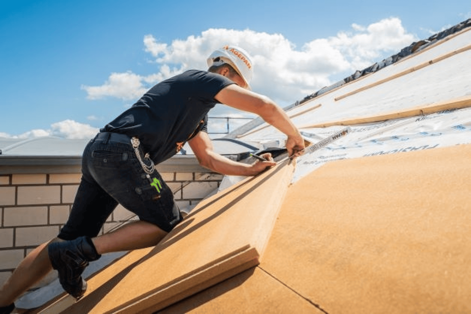 A construction worker wearing a hard hat and shorts is installing insulation panels on a pitched roof in bright sunshine.
