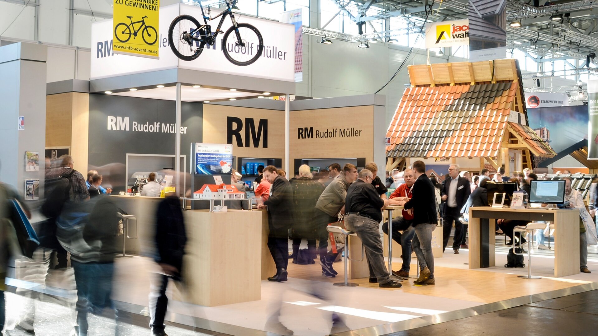 Auf dem Messestand von Rudolf Müller herrscht reges Treiben: Menschen unterhalten sich und stöbern in den Auslagen, ein Fahrrad ist montiert, und ein Modell des Daches steht unter heller Innenbeleuchtung.