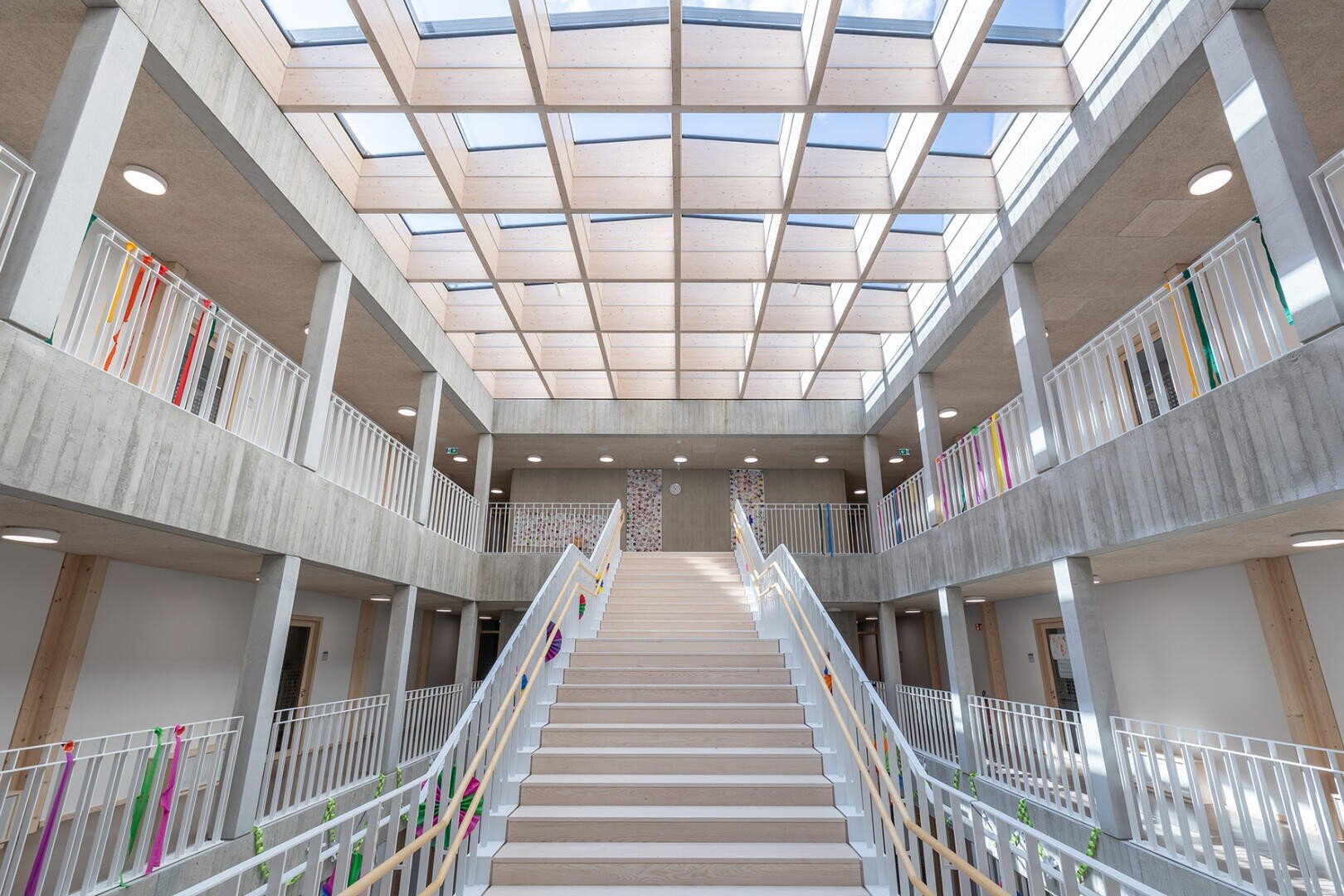 Eine breite Treppe mit weißem Geländer führt zu einem Treppenabsatz in einem hellen, modernen Gebäude. Die Decke ist mit einem großen Raster aus Oberlichtern versehen, die den Raum mit natürlichem Licht füllen. Mehrere Räume mit Türen erstrecken sich über beide Ebenen.