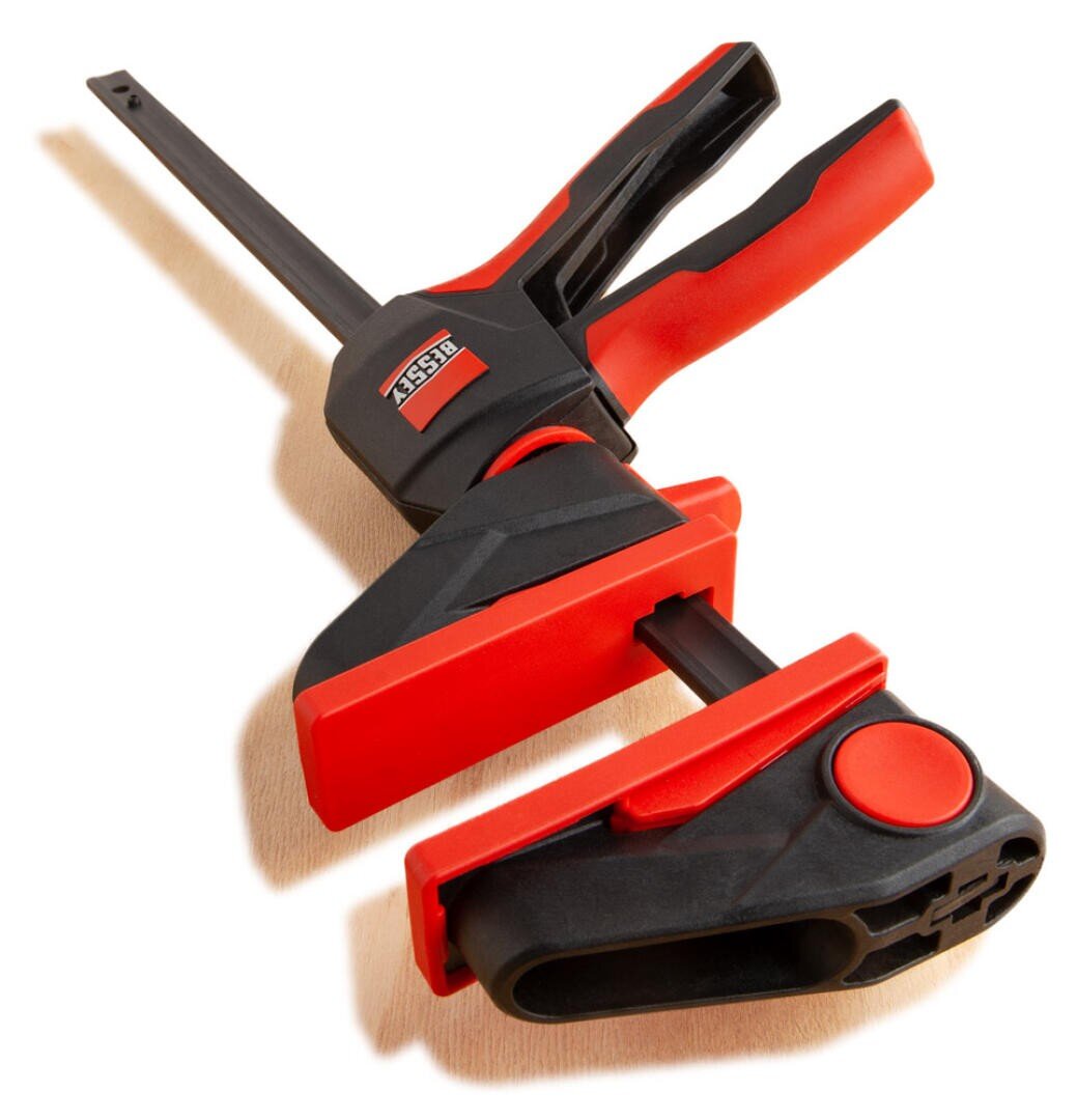 Eine rot-schwarze Stangenklammer mit Auslösegriff, die für die Holzbearbeitung und das Zusammenhalten von Materialien auf einer leichten Unterlage konzipiert ist.