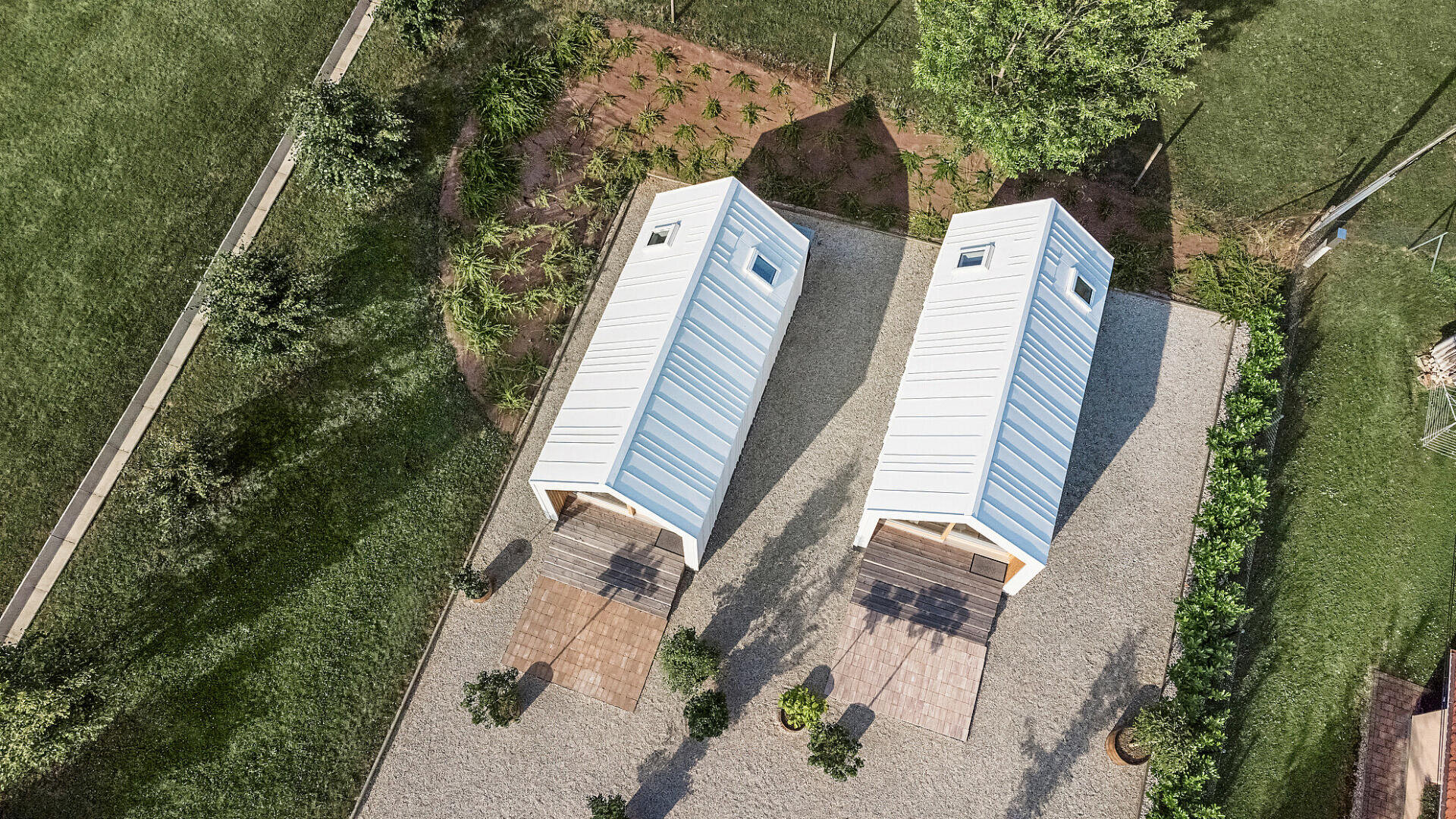 Luftaufnahme von zwei kleinen, weißen, modernen Tiny Houses mit Metalldächern, jedes mit Blick auf einen Terrassenbereich, umgeben von Grünflächen und Kieswegen.