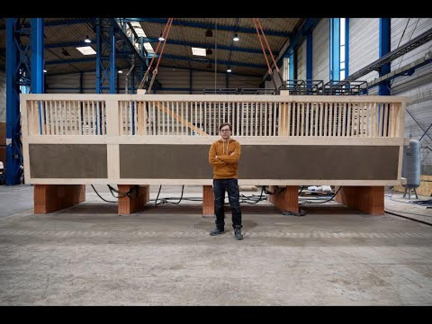 A person stands with folded arms in front of a large, rectangular wooden structure in a spacious industrial hall. The structure consists of a lattice-like upper part and solid panels underneath.
