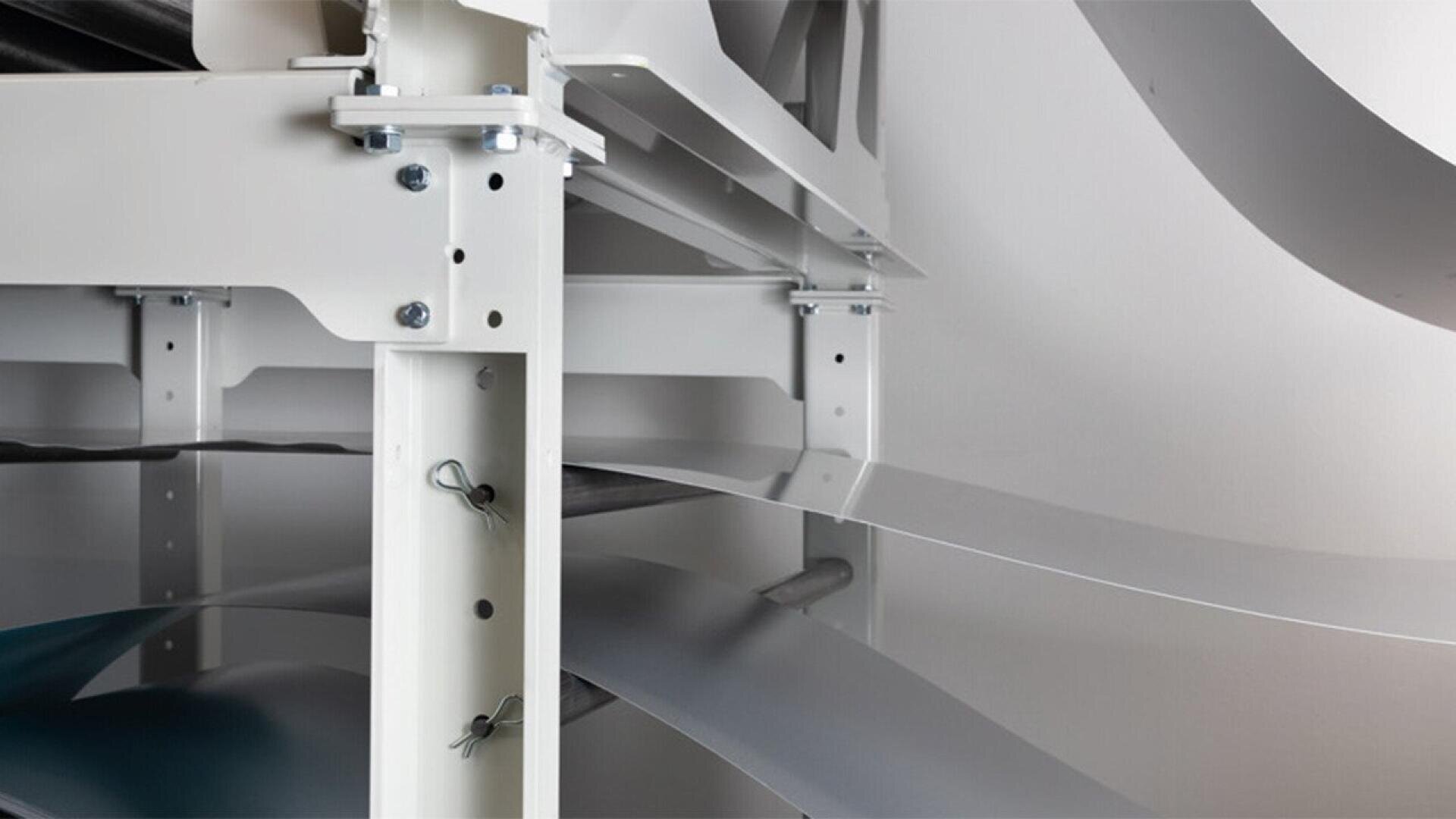 Close-up of a metal industrial shelving unit with sheets of thin metal or plastic material being fed through rollers in a manufacturing or processing facility. The structure is painted light gray or white.
