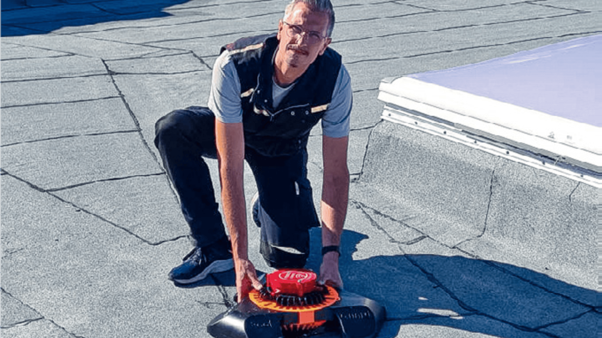 Ein Mann kniet auf einem flachen, grauen Dach und hält eine kleine orange-schwarze Reinigungsmaschine mit der Aufschrift „Ifeex“ in der Hand. Er trägt eine Brille, ein graues Hemd und eine schwarze Weste.