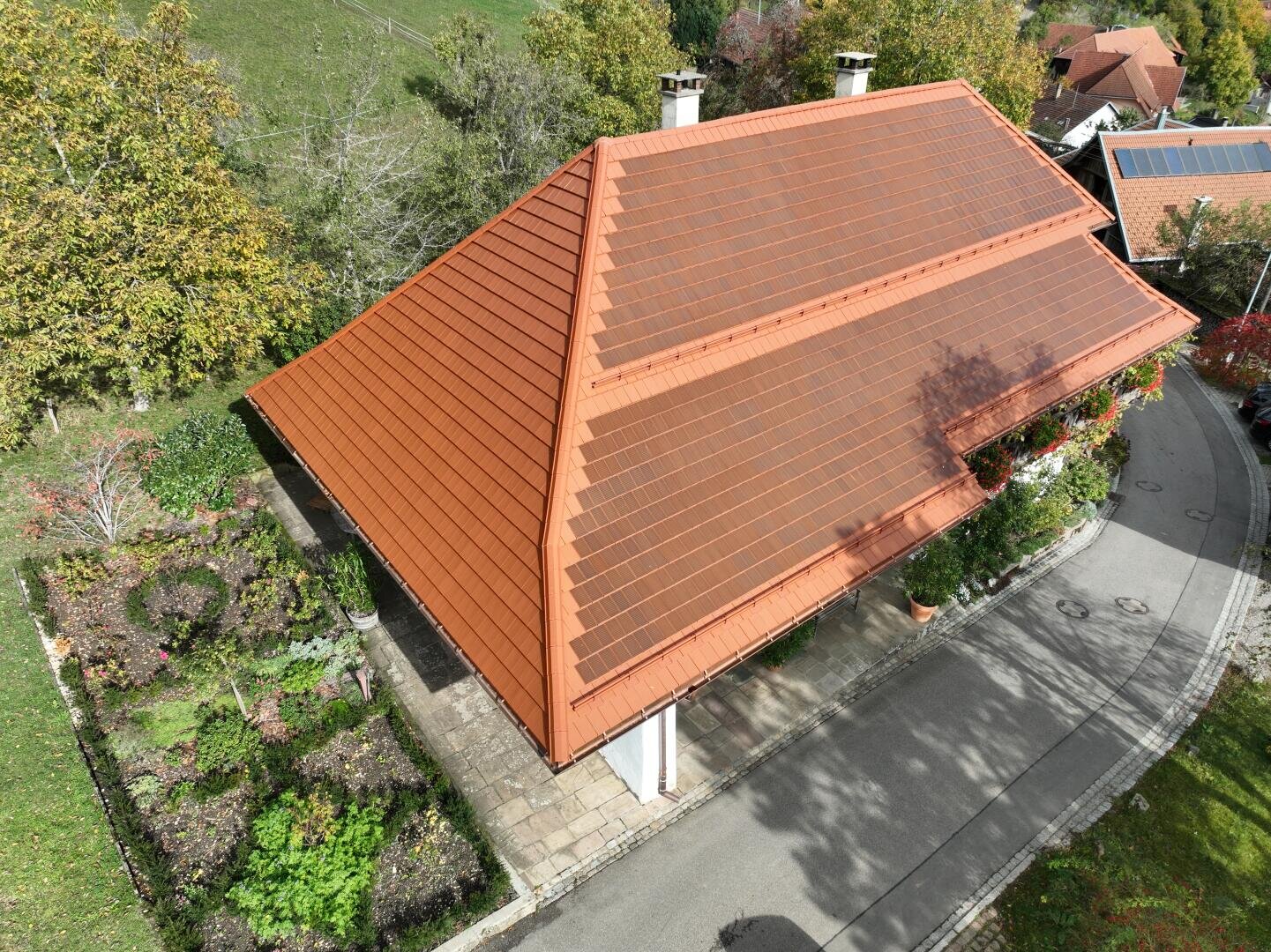 Luftaufnahme eines Hauses mit einem großen roten Ziegeldach mit integrierten Solarmodulen, umgeben von Bäumen, einem Garten und einer geschwungenen Auffahrt.