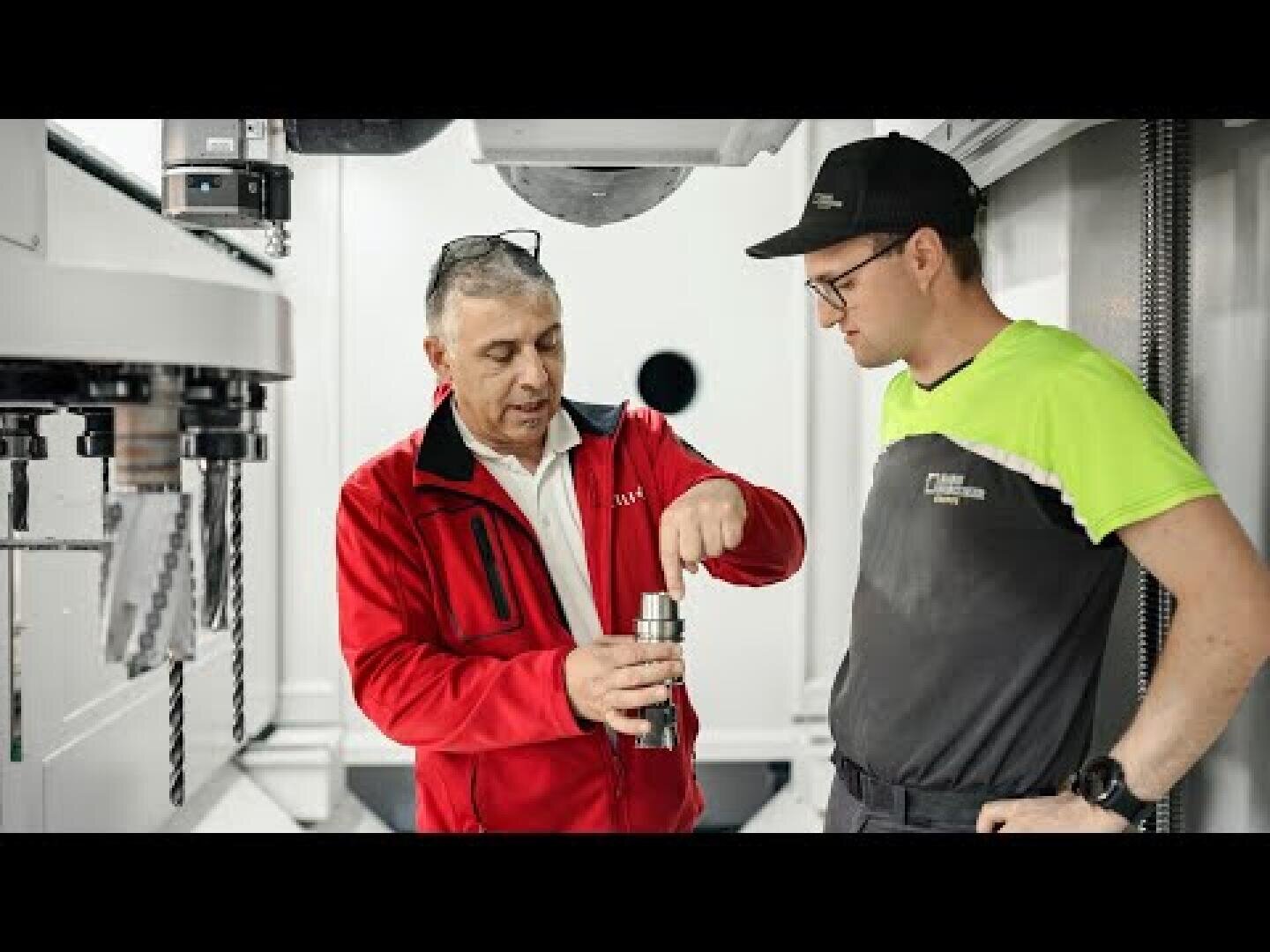 Two men stand in a modern industrial setting; one in a red jacket explains a machine part to the other, who wears a black cap and gray uniform with neon green sleeves. Drills and equipment are visible in the background.