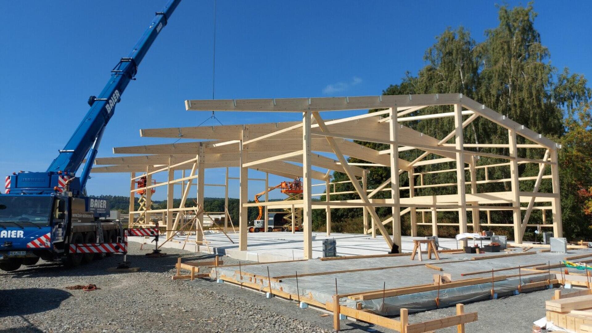 A construction site with a wooden building frame being assembled by cranes and workers under a clear blue sky, surrounded by trees and construction materials.