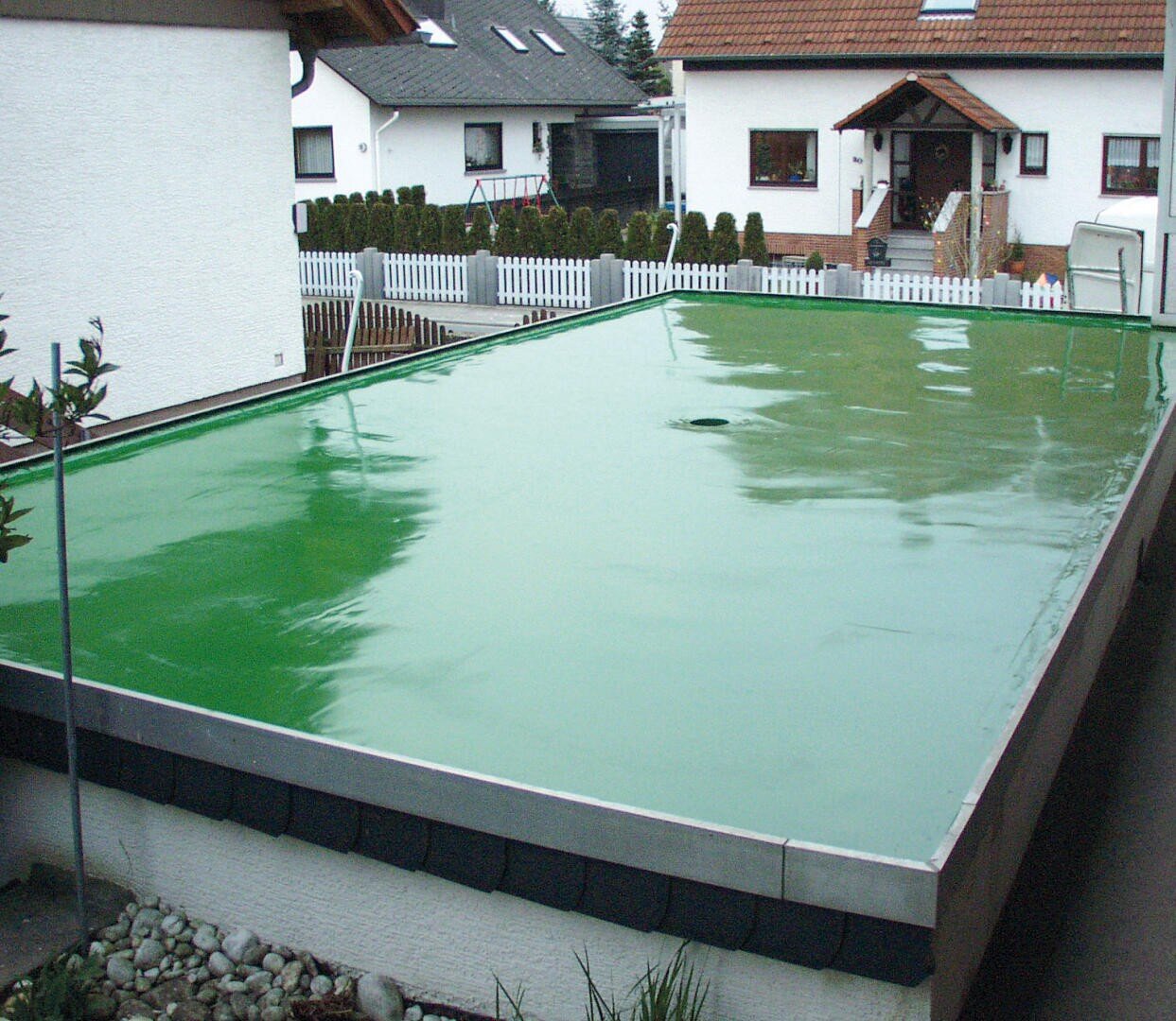 Ein flaches Dach, das mit einer glänzenden grünen, wasserdichten Membran bedeckt ist, liegt zwischen Wohnhäusern mit Gärten und weißen Zäunen in einem Vorstadtviertel.