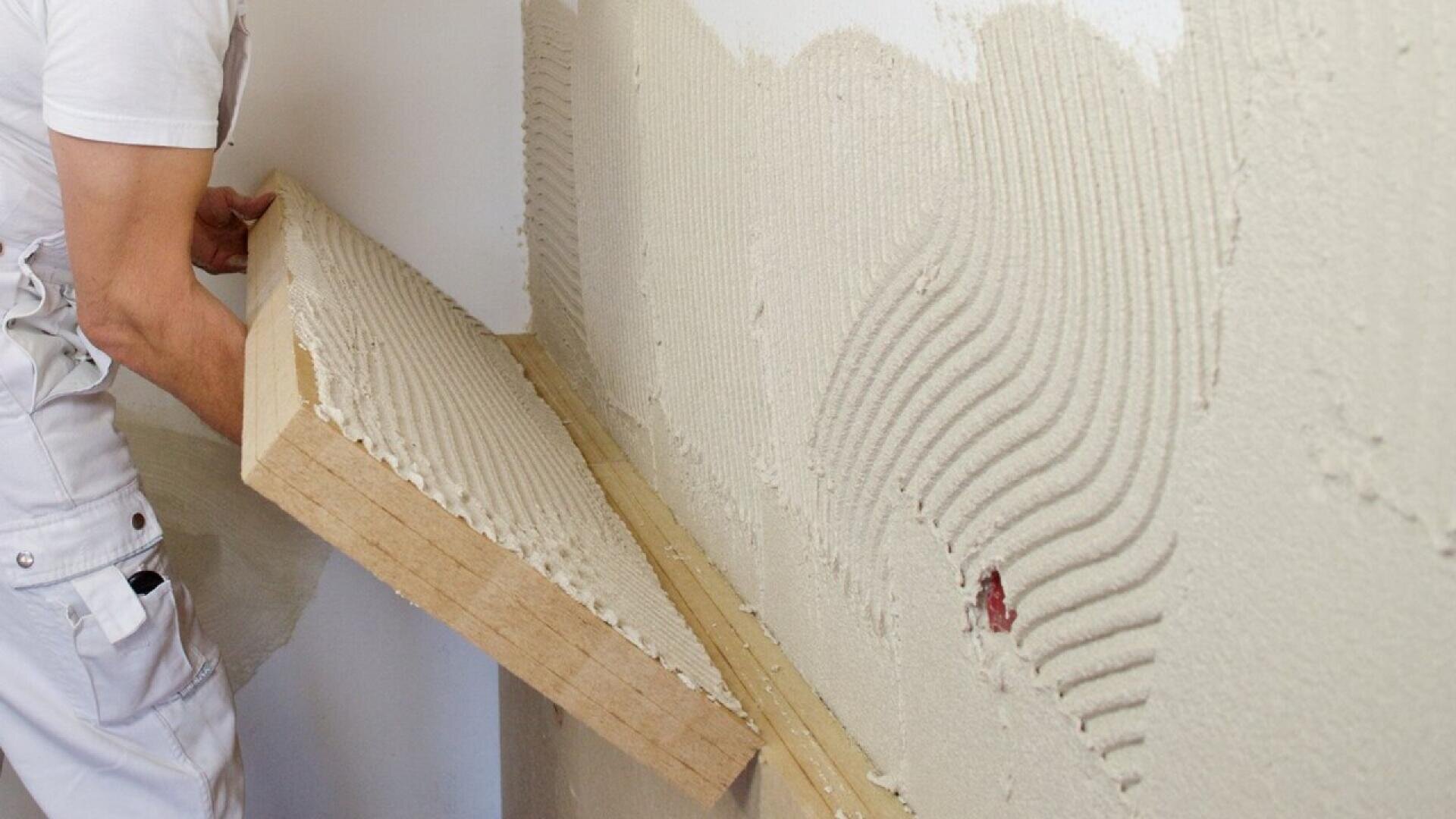 A person applies insulation board to a wall with grooves of adhesive, wearing white work clothes and holding the board at an angle, ready to attach it to the wall.