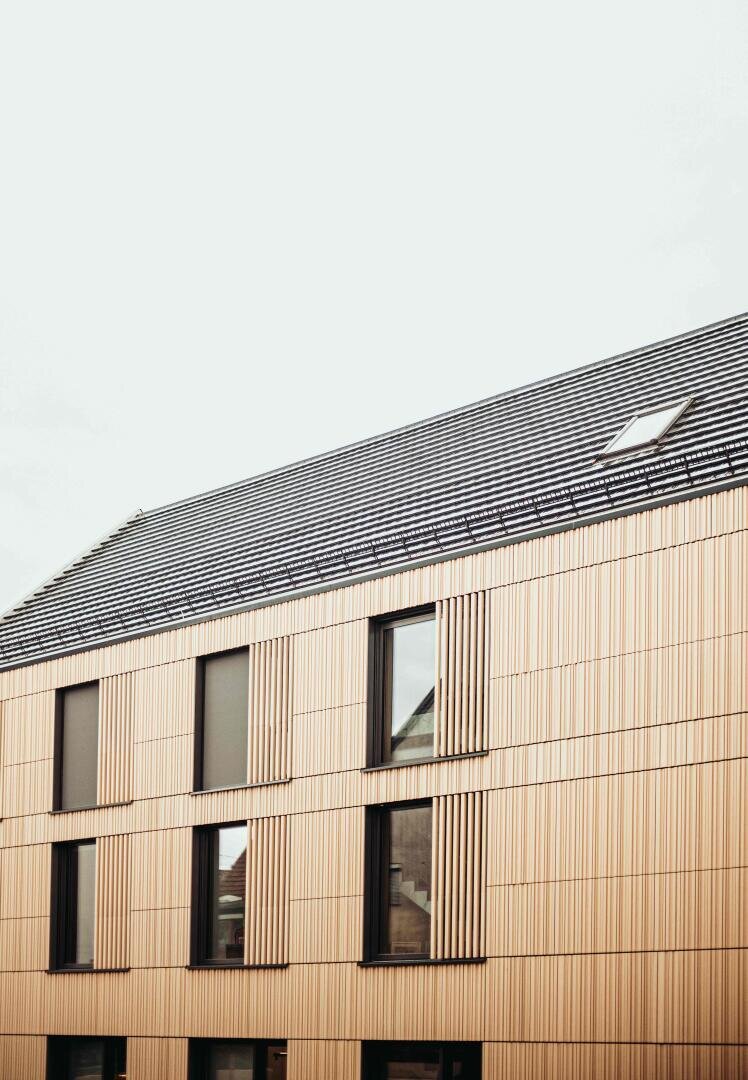 A modern building with a sloping, dark gray roof and light-colored wood cladding. Large, rectangular windows are evenly spaced on the façade under an overcast sky.