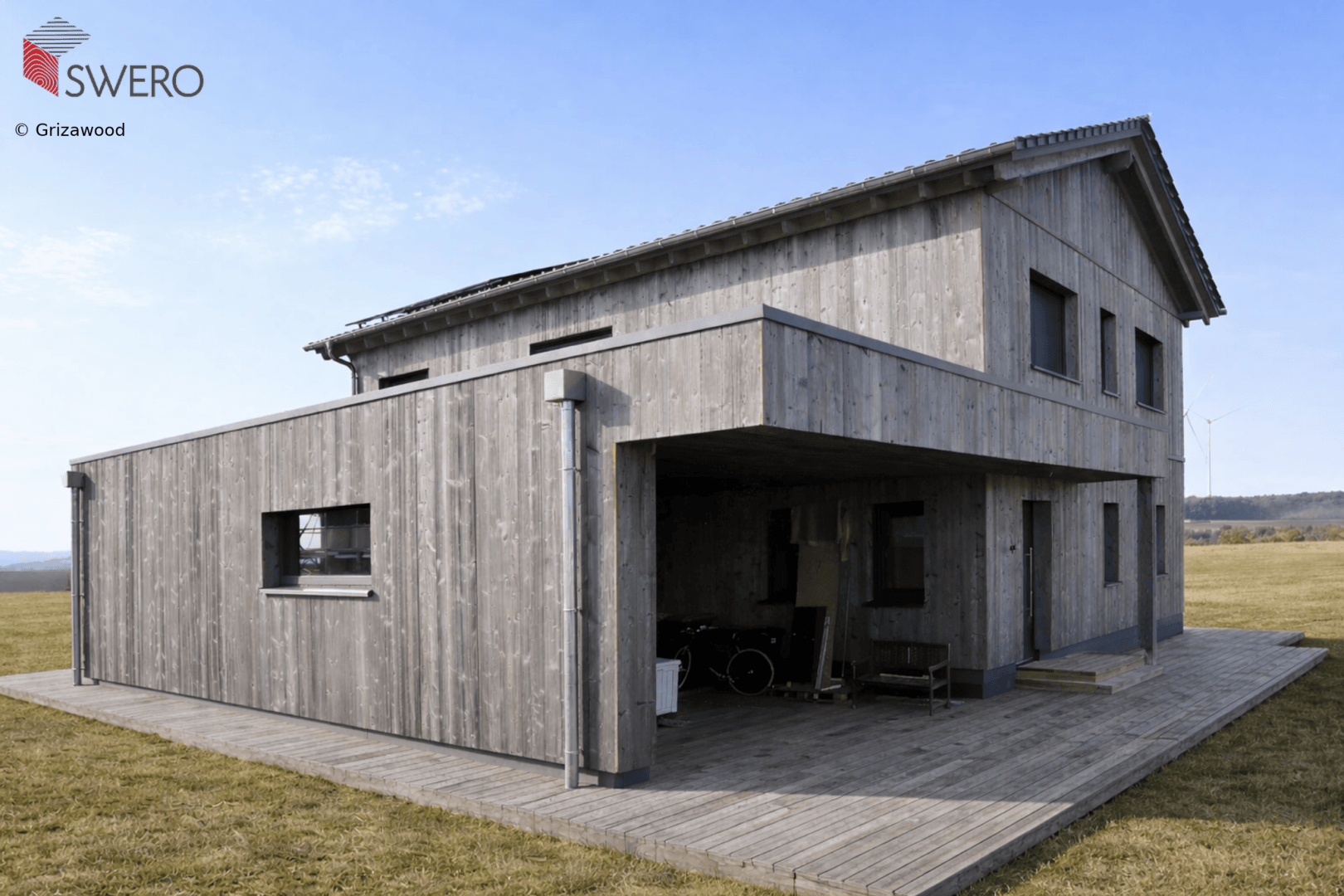 Modernes zweistöckiges Haus aus grauem Holz mit einem Flachdachanbau und einer überdachten Veranda, gelegen auf einer offenen Wiese unter einem teilweise bewölkten Himmel.