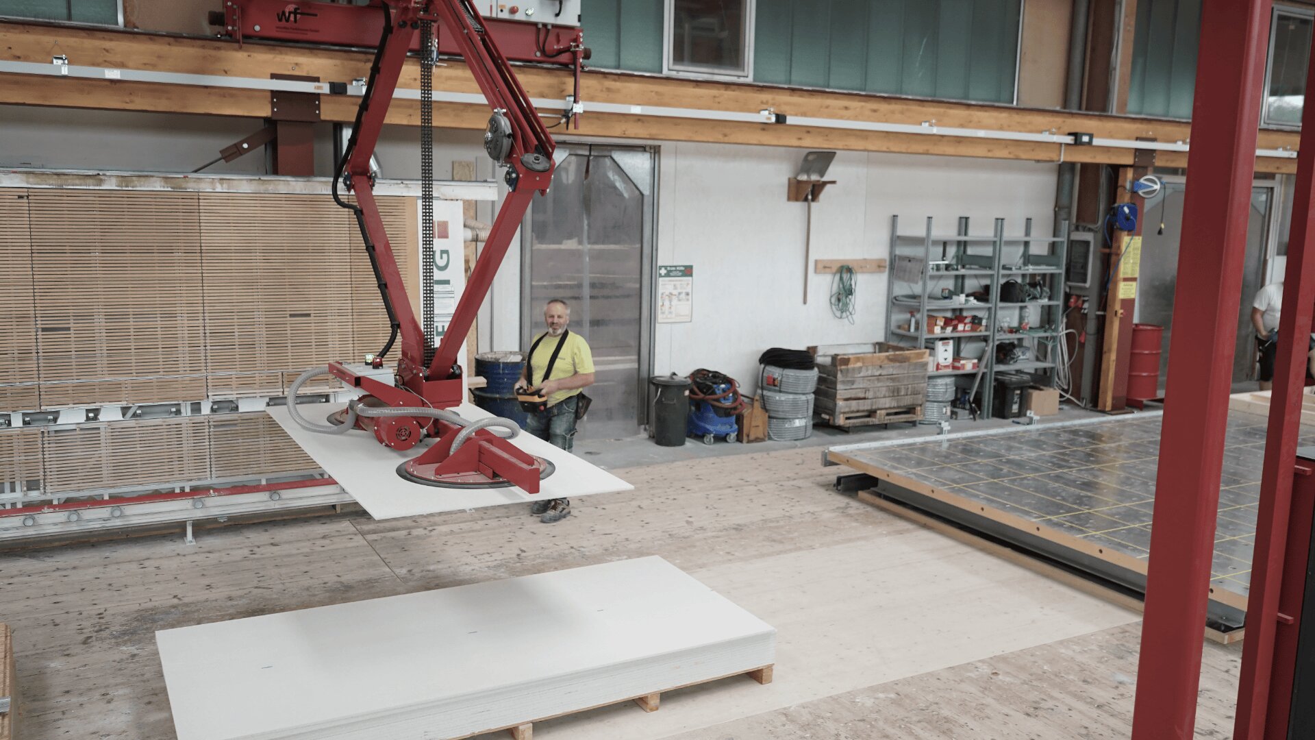 Ein Arbeiter in einem gelben Hemd bedient einen roten Roboterarm, der in einer geräumigen Industriewerkstatt mit Regalen, Geräten und Holzbalken eine große weiße Platte anhebt.