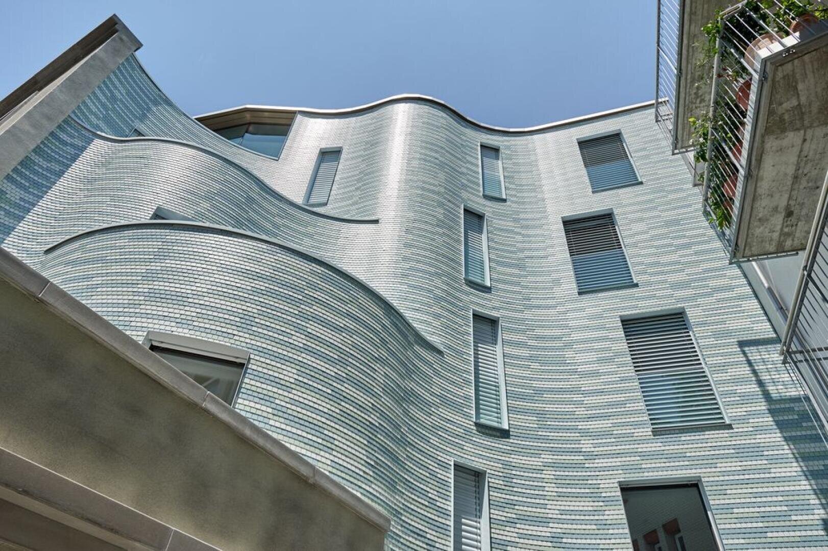 Ein modernes Gebäude mit einer gewellten, mit hellblauen Kacheln bedeckten Fassade mit mehreren rechteckigen Fenstern, von unten gesehen vor einem klaren Himmel. Auf der rechten Seite ist ein Balkon mit Pflanzen zu sehen.