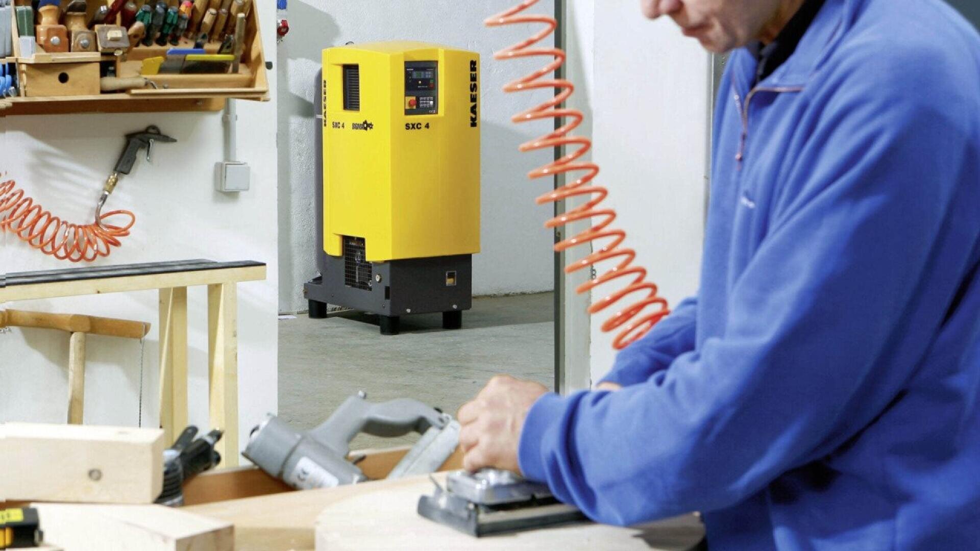 A person in a blue jacket uses a power tool on a wooden surface in a workshop. In the background, a yellow industrial machine and various woodworking tools are visible on shelves and walls.