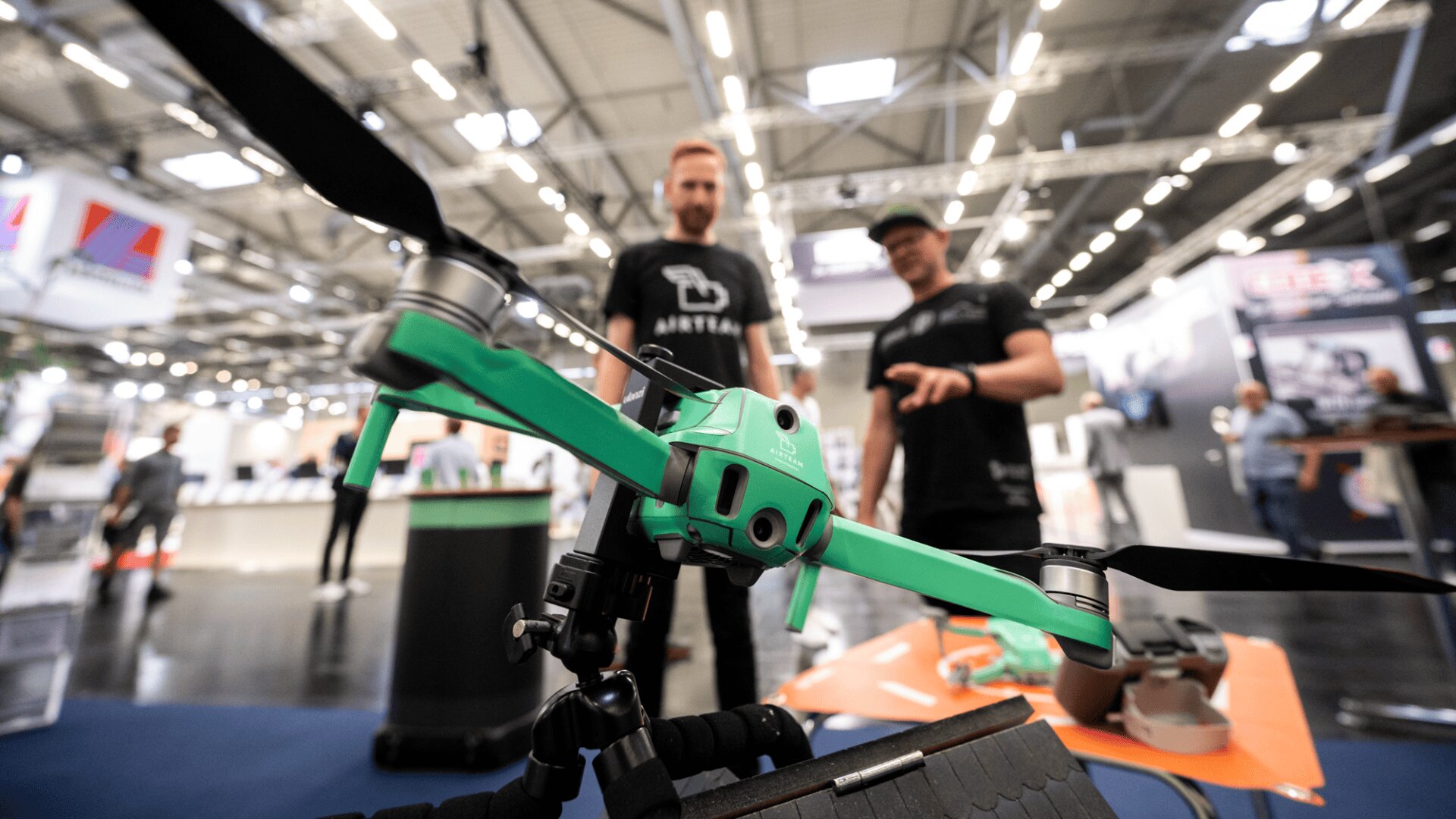 A green drone is displayed in focus at an indoor tech expo, with two people in black shirts standing in the blurred background examining it. Bright lights and booths are visible throughout the space.