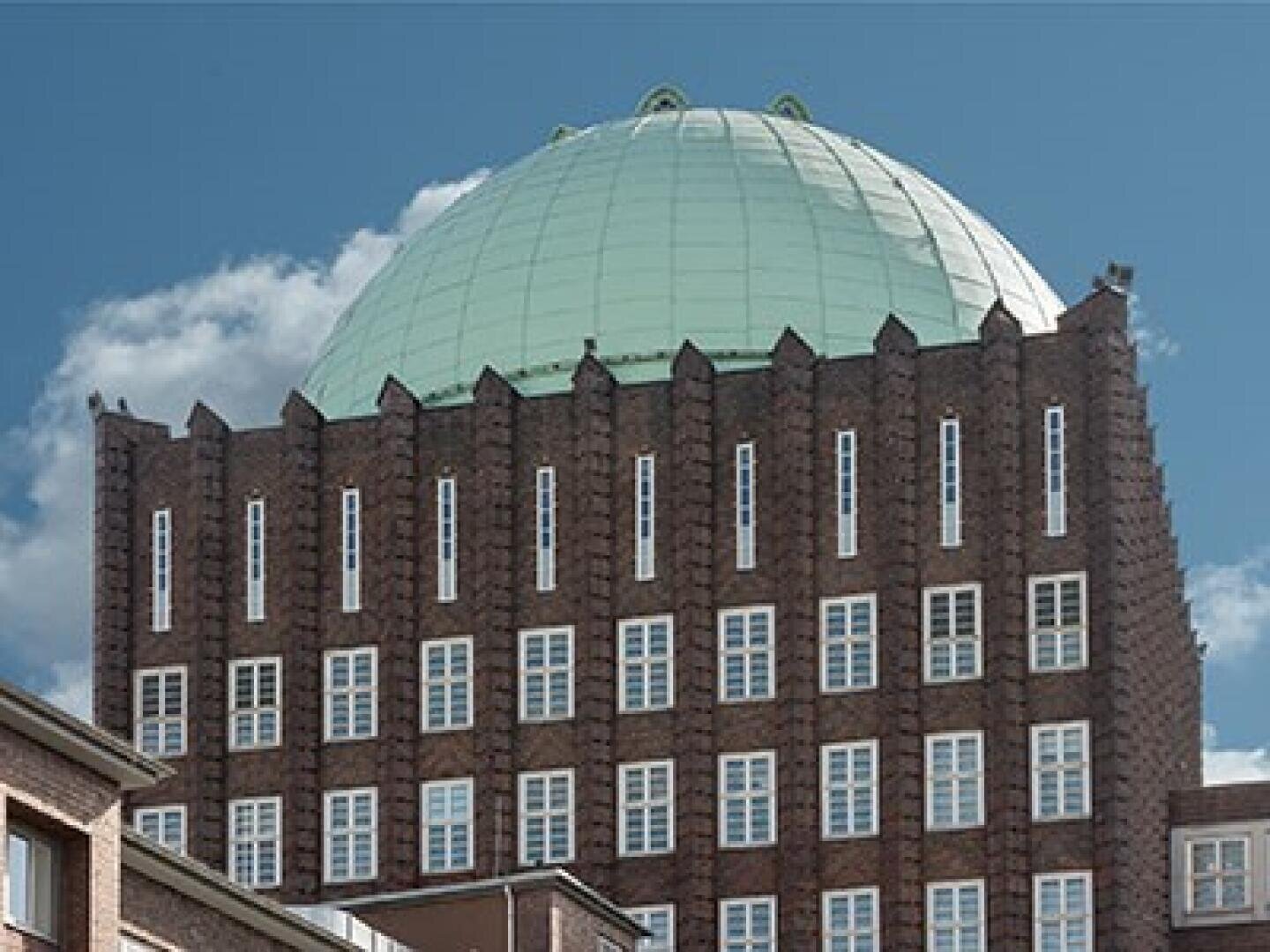 Vor einem blauen Himmel mit vereinzelten Wolken steht ein hohes Backsteingebäude mit vielen rechteckigen Fenstern und einem großen, grünen Kuppeldach.