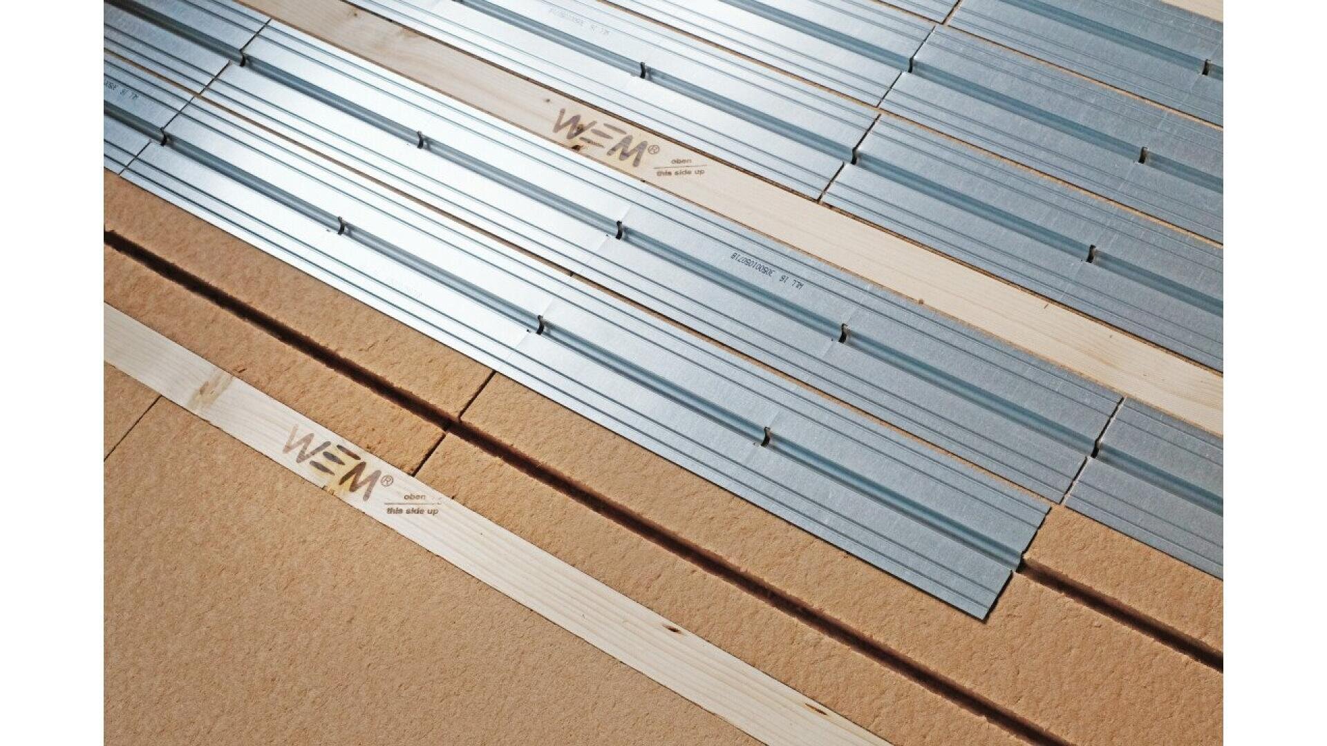Close-up of wooden planks and metal rails arranged in parallel rows on a surface, with some pieces labeled “WEM.” The image shows construction materials, possibly for flooring or wall installation.