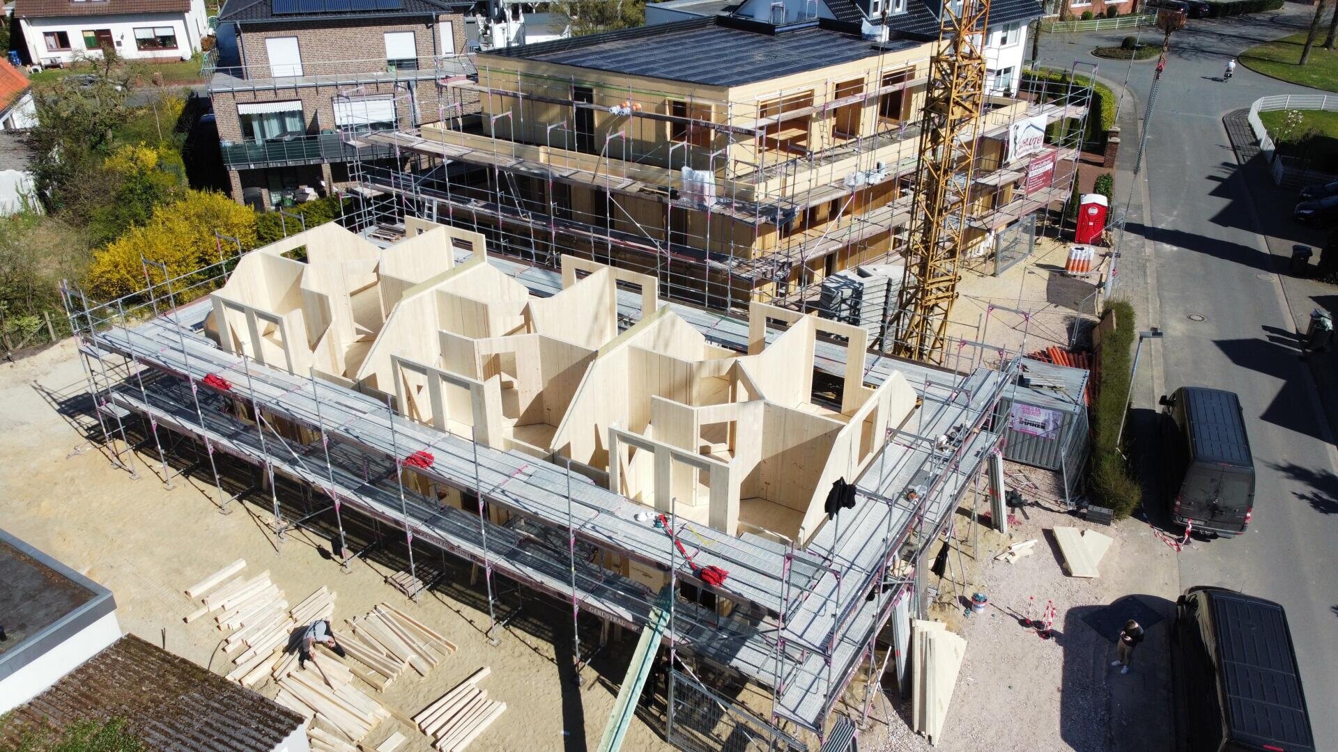 Luftaufnahme einer Baustelle mit einem Gebäude aus Holzpaneelen im Bau. Das Bauwerk ist von Gerüsten umgeben, und man sieht Arbeiter, Material und Ausrüstung. Ein angrenzendes Gebäude scheint weiter im Bau zu sein.