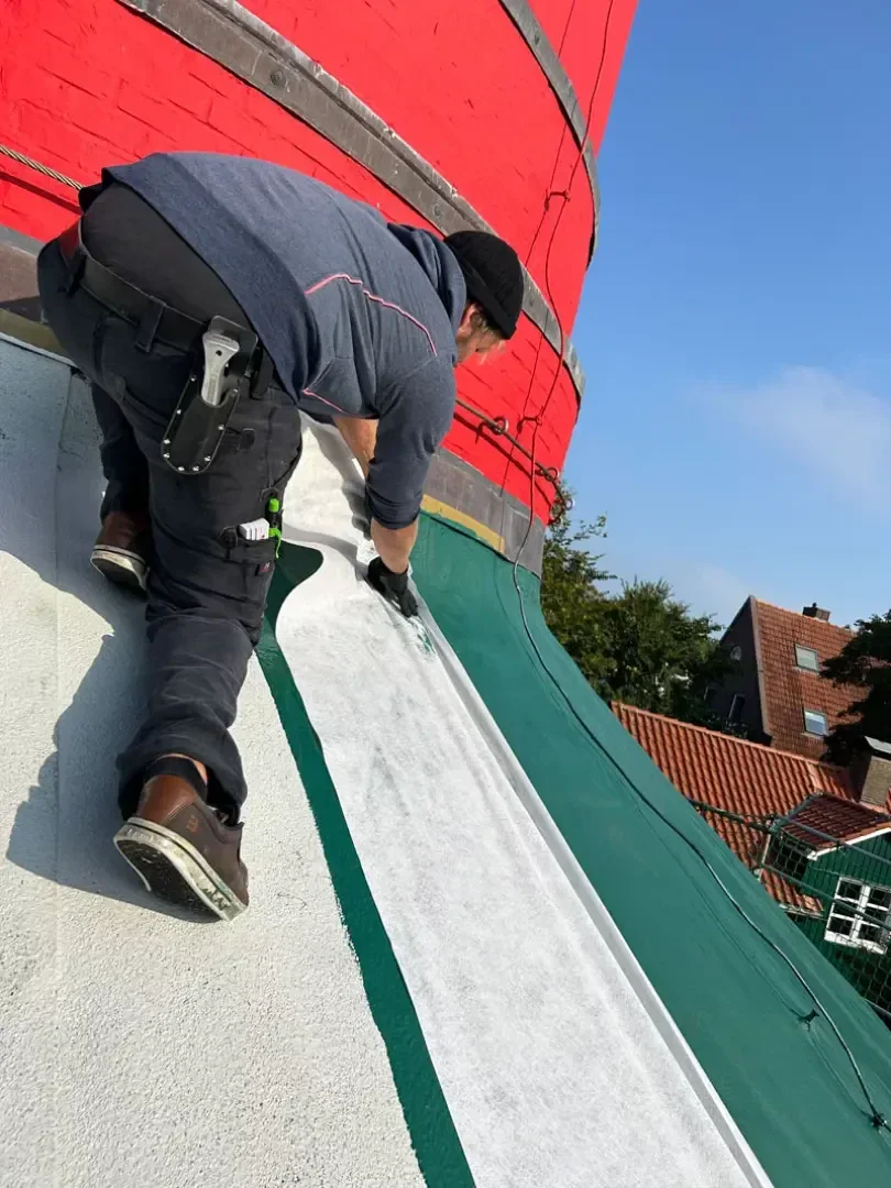 Ein Arbeiter installiert oder befestigt Material auf dem Schrägdach eines Gebäudes. Unter einem klaren blauen Himmel sind rote und grüne Flächen sichtbar. Im Hintergrund sind Dächer und Bäume zu sehen.
