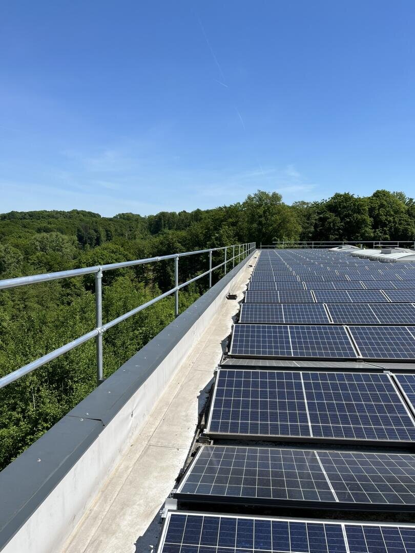 Reihen von Solarzellen auf einem Dach fangen das Sonnenlicht unter einem klaren blauen Himmel ein, umgeben von üppig grünen Bäumen und bewaldeten Hügeln im Hintergrund.