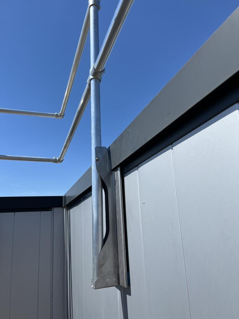 Eine Nahaufnahme eines Metallgeländers, das an der oberen Ecke eines grauen Metallzauns oder -tors befestigt ist, mit einem klaren blauen Himmel im Hintergrund. Das Bild unterstreicht die Struktur und Verbindung der Metallteile.