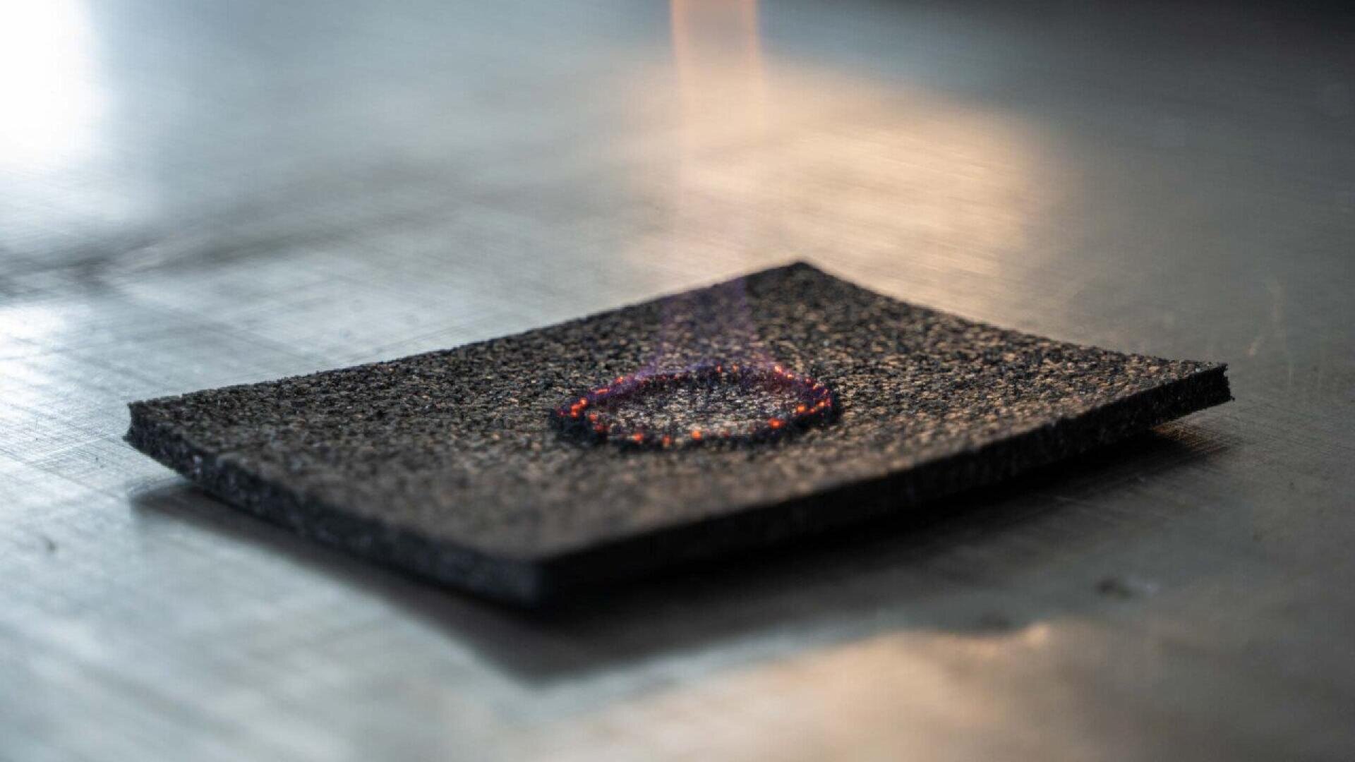 A close-up of a rectangular black material with a smoldering, glowing red ring burned into its center, placed on a metallic surface. Thin wisps of smoke or heat rise from the burned area.