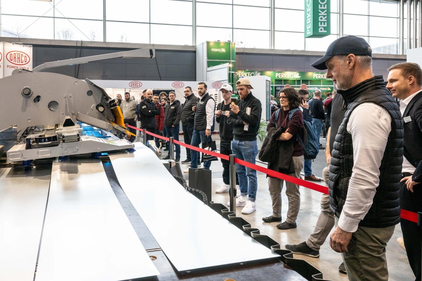 Eine Gruppe von Personen beobachtet eine große Industriemaschine, die in einer Halle in Betrieb ist, wobei die Beobachter durch rote Barrieren von der Anlage getrennt sind. Einige Personen machen Fotos oder Videos.