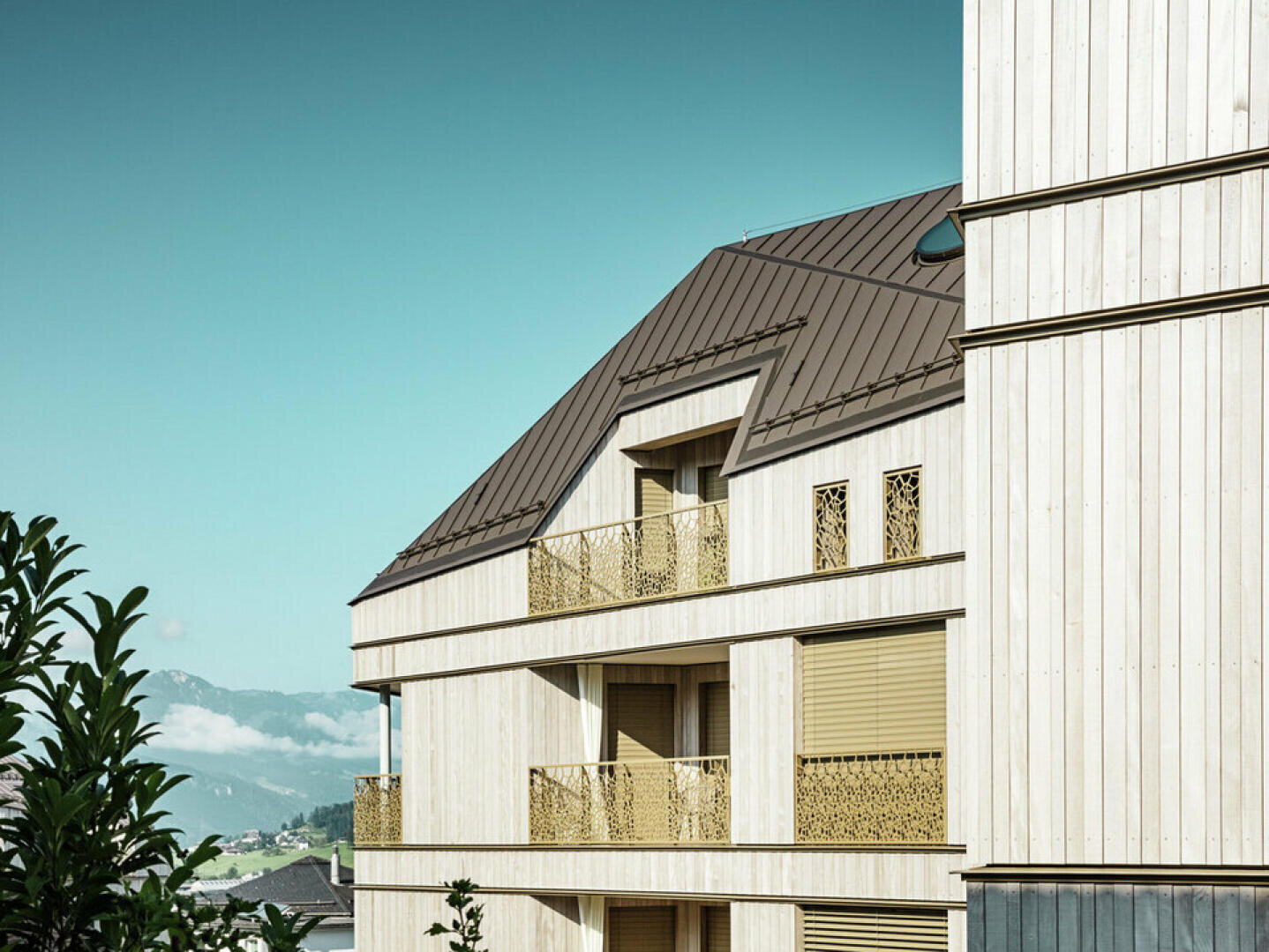 Modernes Holzgebäude mit goldenem Geländer und Fensterläden sowie einem schrägen dunklen Dach. Im Hintergrund sind ein klarer blauer Himmel und ferne Berge zu sehen, im Vordergrund etwas Grün.