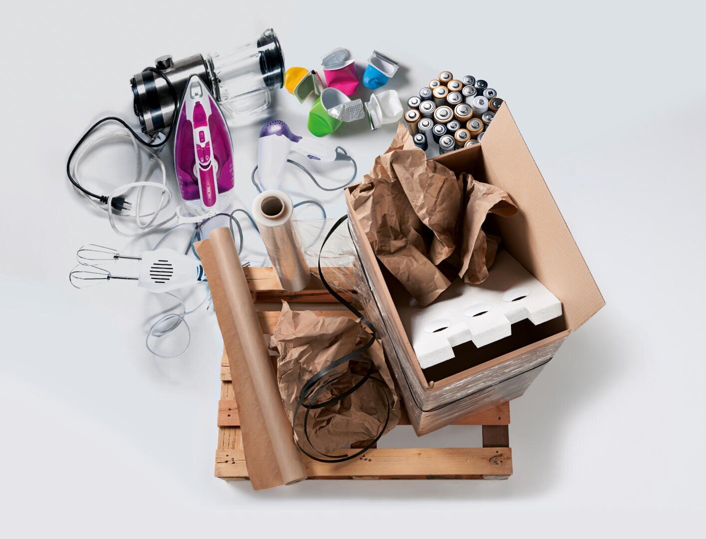 On a wooden pallet is a cardboard box filled with brown paper, foam, plastic film and cables. Next to it are batteries, small appliances, paint cans and a purple iron on a light gray background.