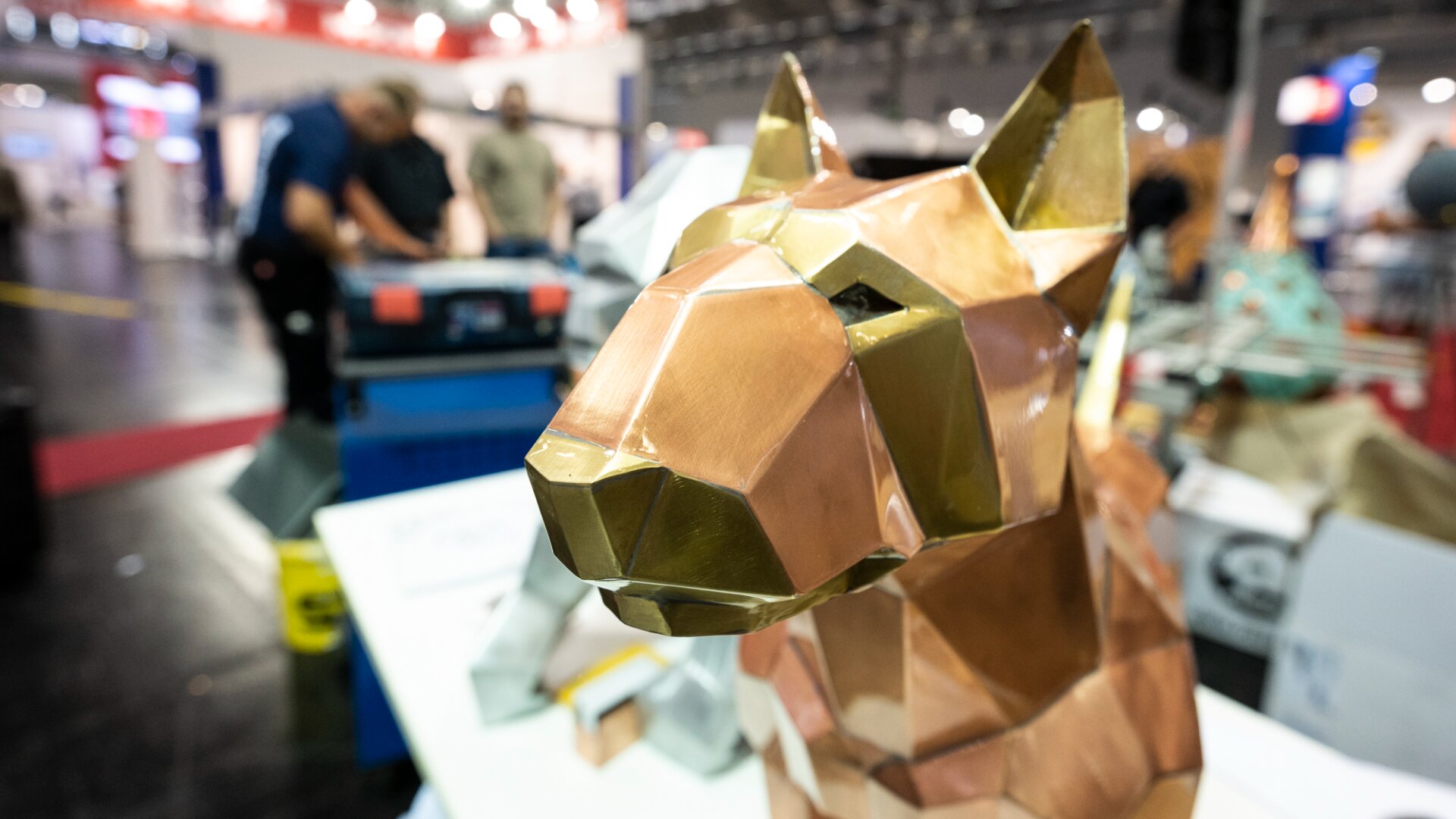 Eine geometrische, metallische Skulptur eines Hundekopfes steht auf einem Tisch bei einer Indoor-Veranstaltung, und im Hintergrund sind unscharfe Menschen und Geräte zu sehen.