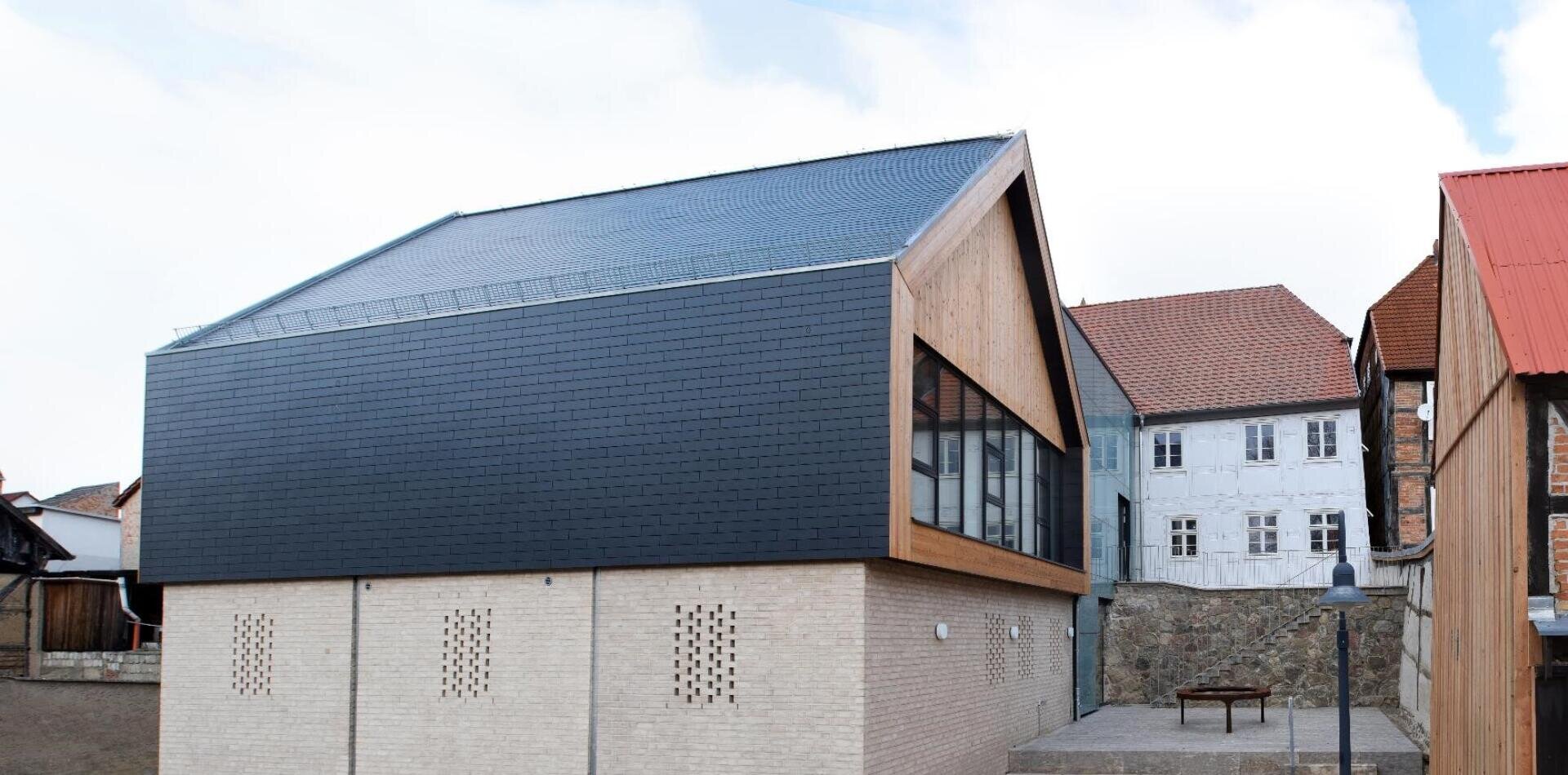 Modern building with a beige brick basement and a black, sloping upper façade. Large windows line the side and it is surrounded by older houses with red roofs. There is a stone wall and a small courtyard in the foreground.