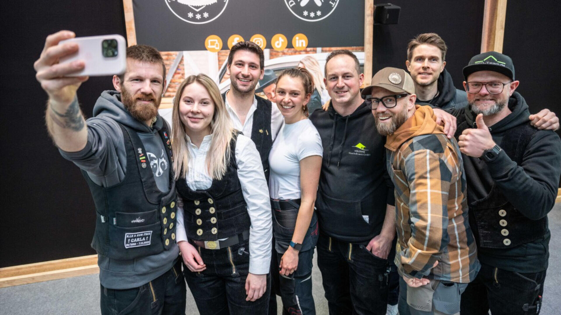 Eine Gruppe von acht lächelnden Menschen, einige in Arbeitswesten und Freizeitkleidung, posiert gemeinsam für ein Selfie in einem Innenbereich vor einem dunklen Hintergrund mit weißen Logos und Social-Media-Symbolen.
