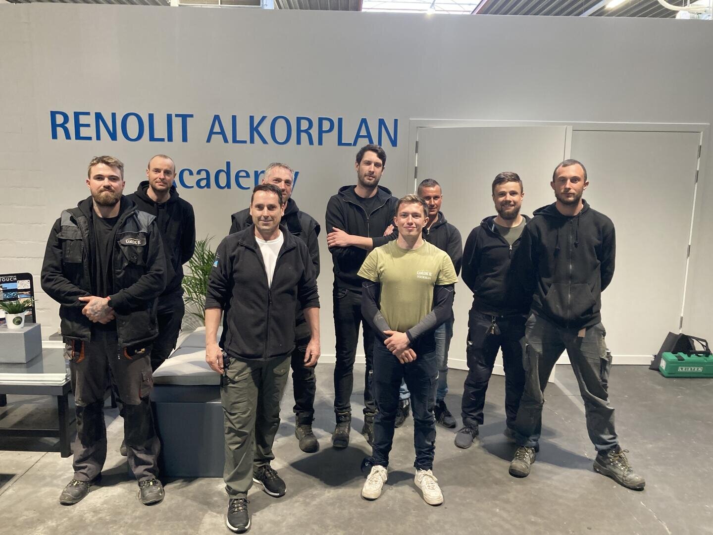 Neun Männer stehen und posieren für ein Gruppenfoto in einem Gebäude mit weißen Wänden. Im Hintergrund ist ein Schild mit der Aufschrift RENOLIT ALKORPLAN academy zu sehen. Sie sind in legerer Arbeitskleidung gekleidet.