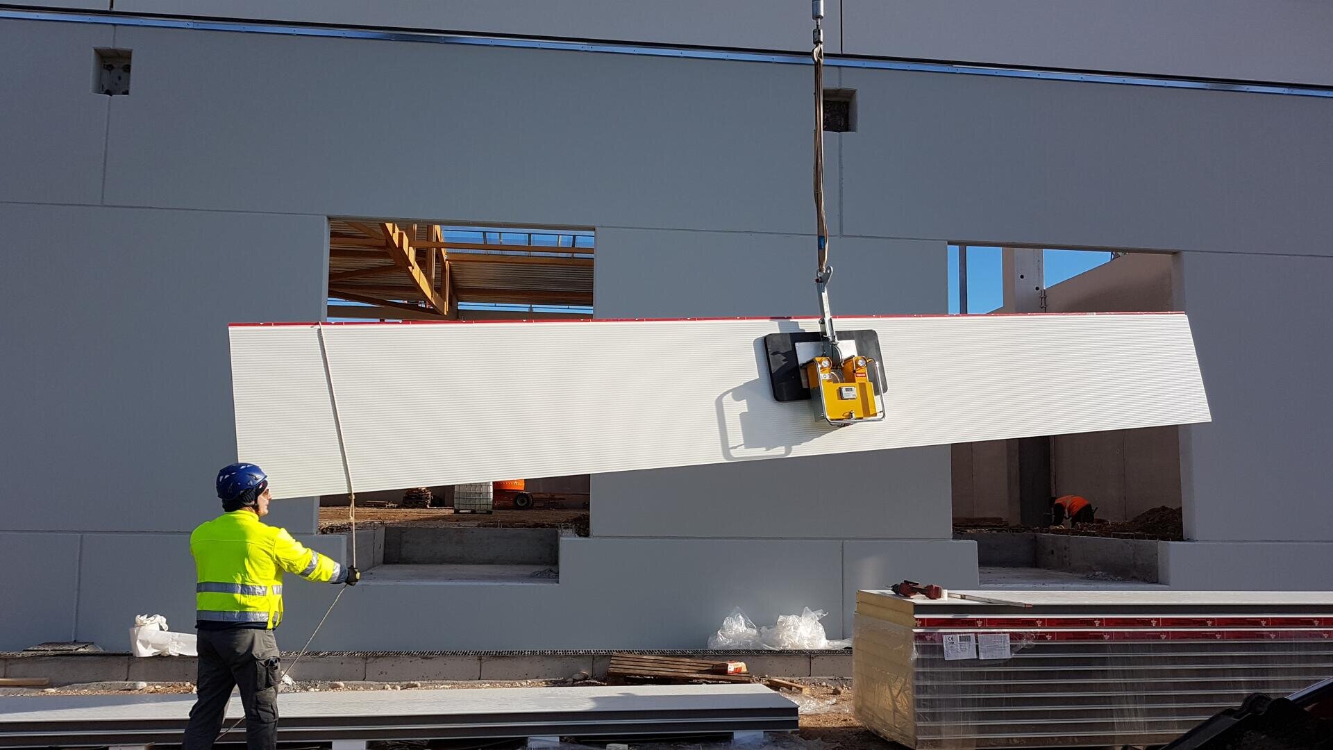 A construction worker in a yellow jacket and blue helmet is guiding a large white wall panel being lifted by a crane in front of a partially constructed building.