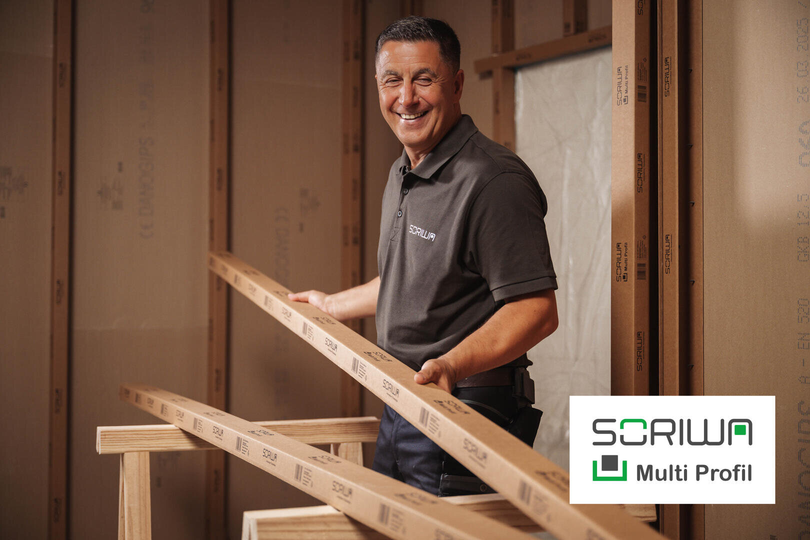 Ein lächelnder Mann in einem SORIWA Polohemd hält zwei Holzbretter in einem im Bau befindlichen Raum. Das SORIWA Multi Profil Logo erscheint in der unteren rechten Ecke.