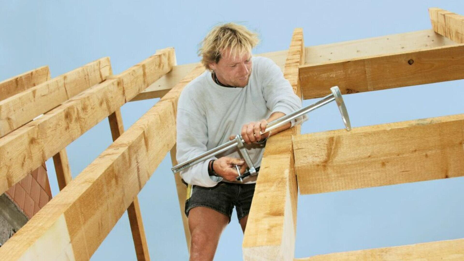 A person wearing a light sweatshirt and shorts is working on a wooden roof frame, using a metal tool to secure large beams against a clear blue sky.