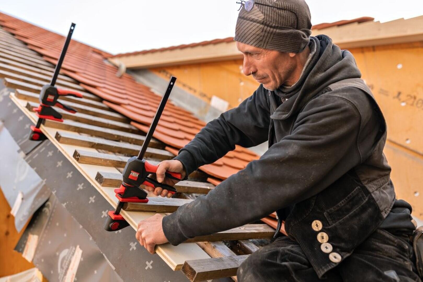 Ein Arbeiter mit Mütze und Latzhose befestigt mit Klemmen Holzbretter auf einem schrägen, im Bau befindlichen Dach, wobei im Hintergrund rote Dachziegel und Holzbalken zu sehen sind.