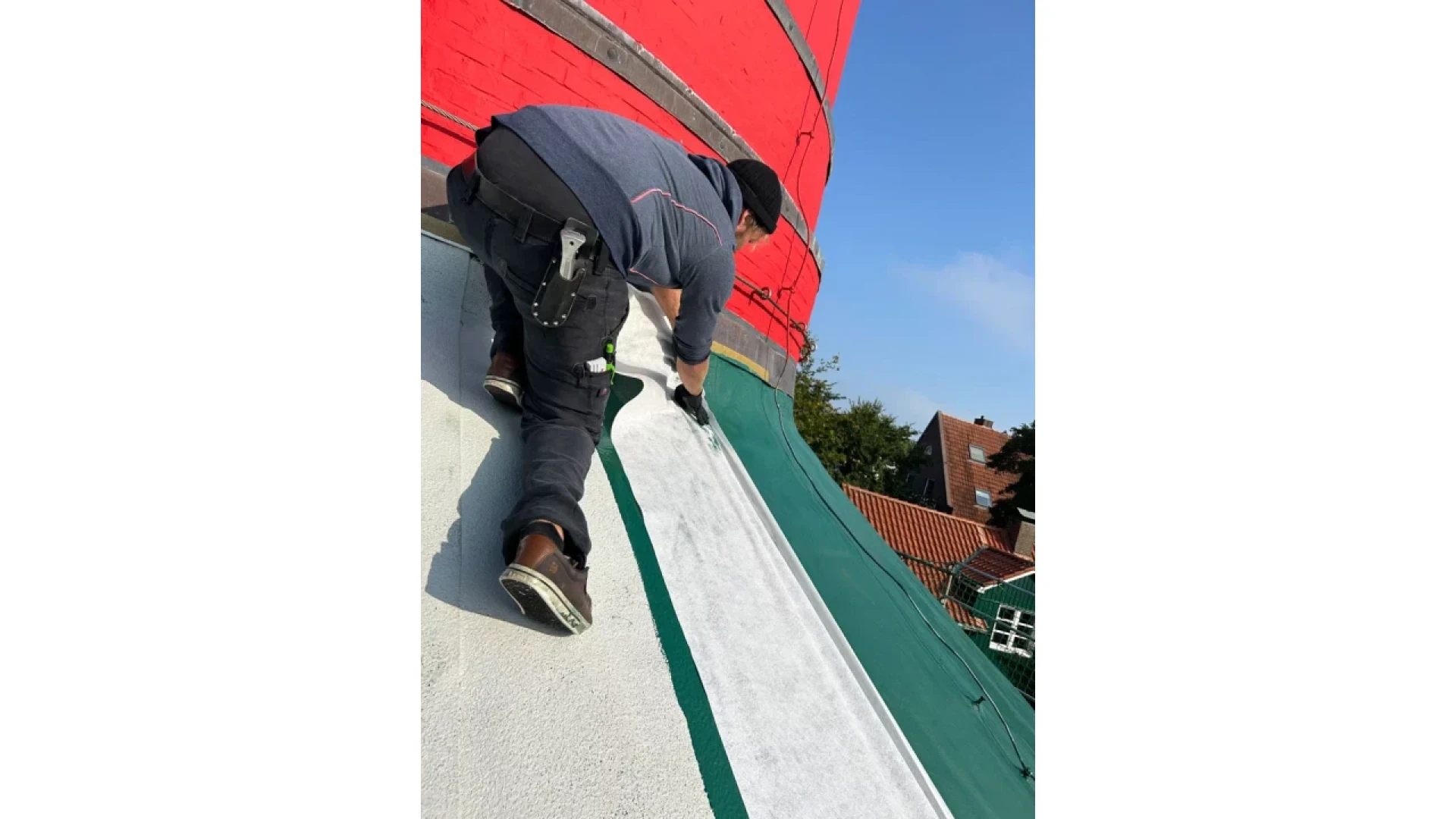 A worker in a gray shirt and black pants applies material to a sloped rooftop next to a red brick building, with houses and trees visible in the background under a clear blue sky.