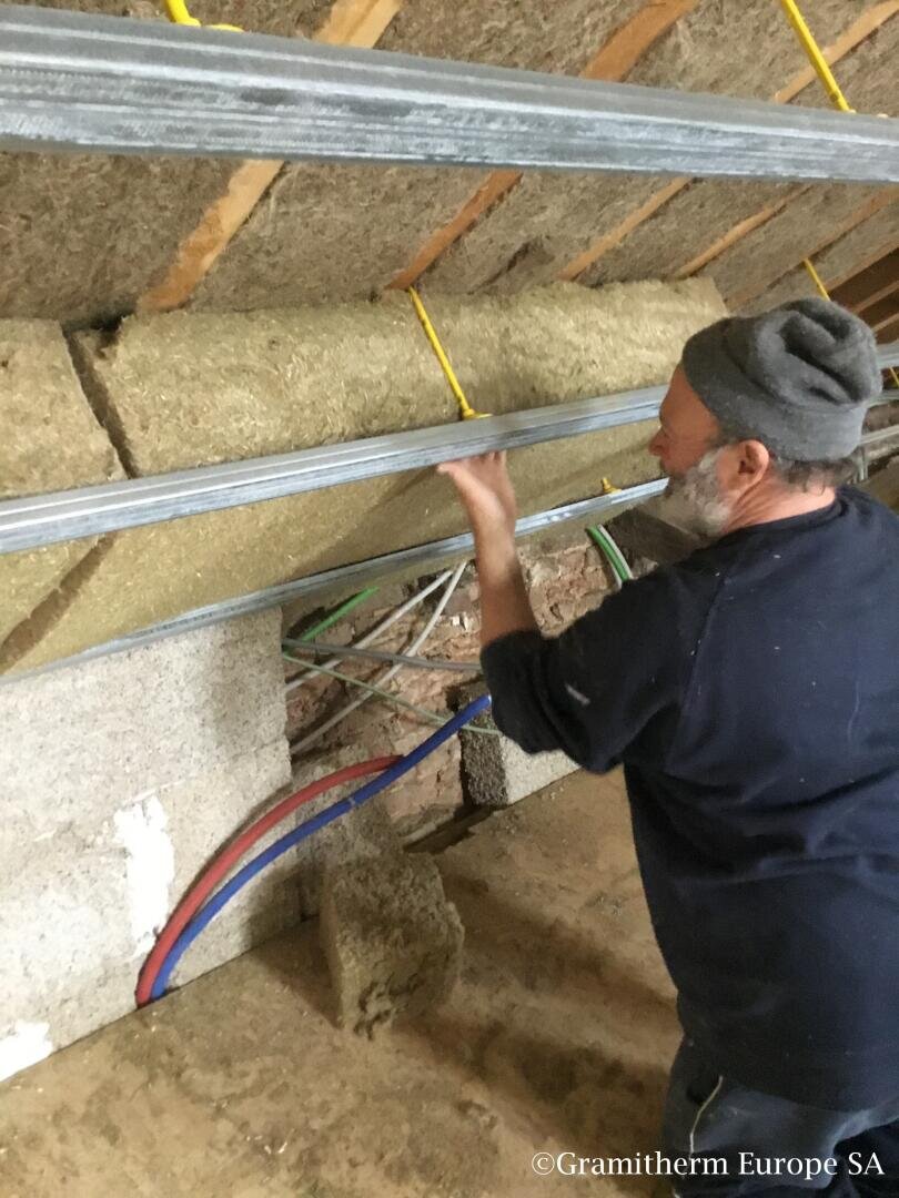 Ein Mann mit Beanie und dunkler Kleidung installiert Naturfaser-Dämmplatten und Metallrahmen in einem im Bau befindlichen Gebäude. Freiliegende Rohre und gelbe Stromkabel sind sichtbar.