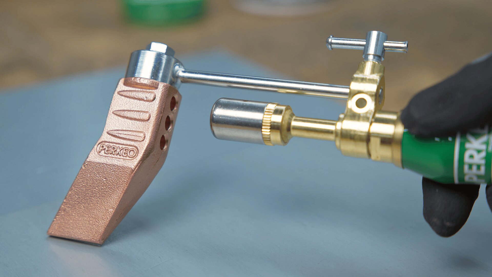 Eine behandschuhte Hand hält eine Lötlampe aus Messing und erhitzt eine Lötvorrichtung aus Kupfer mit Belüftungslöchern und Perko-Branding, die auf einer Metallfläche steht.