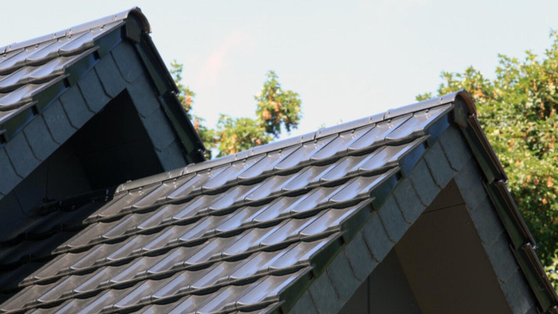 Close-up view of two sloped rooftops with dark gray, shiny, overlapping roof tiles, set against a background of green trees and blue sky.