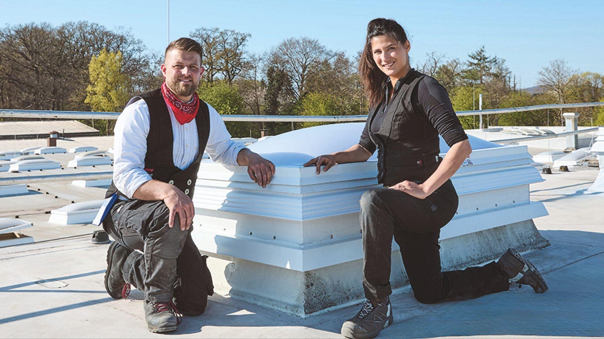 Two people in work clothes and boots kneel on a flat roof next to a white skylight construction, with trees and a clear blue sky in the background.