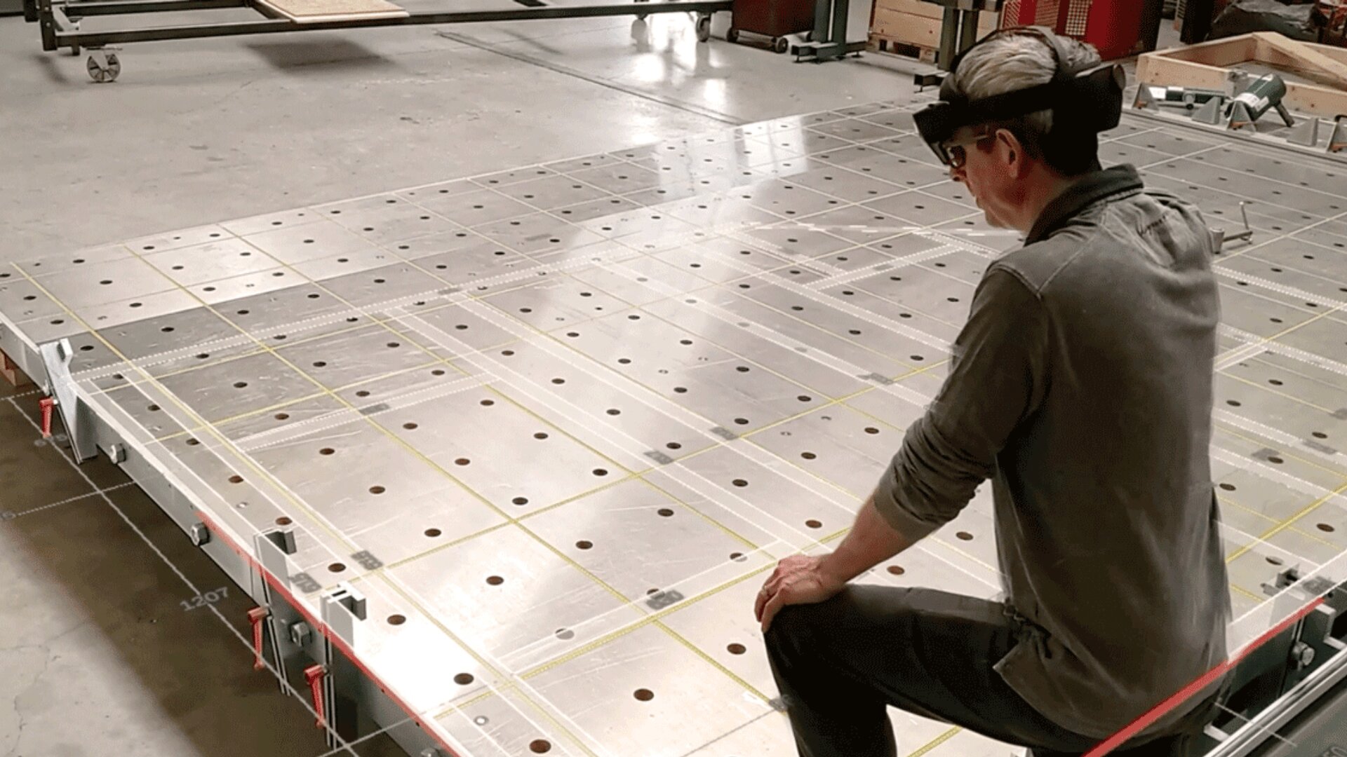 Eine Person mit Augmented-Reality-Brille hockt in einer Industriewerkstatt neben einem großen Metallarbeitstisch mit einem Raster aus Löchern und sich kreuzenden gelben Linien.